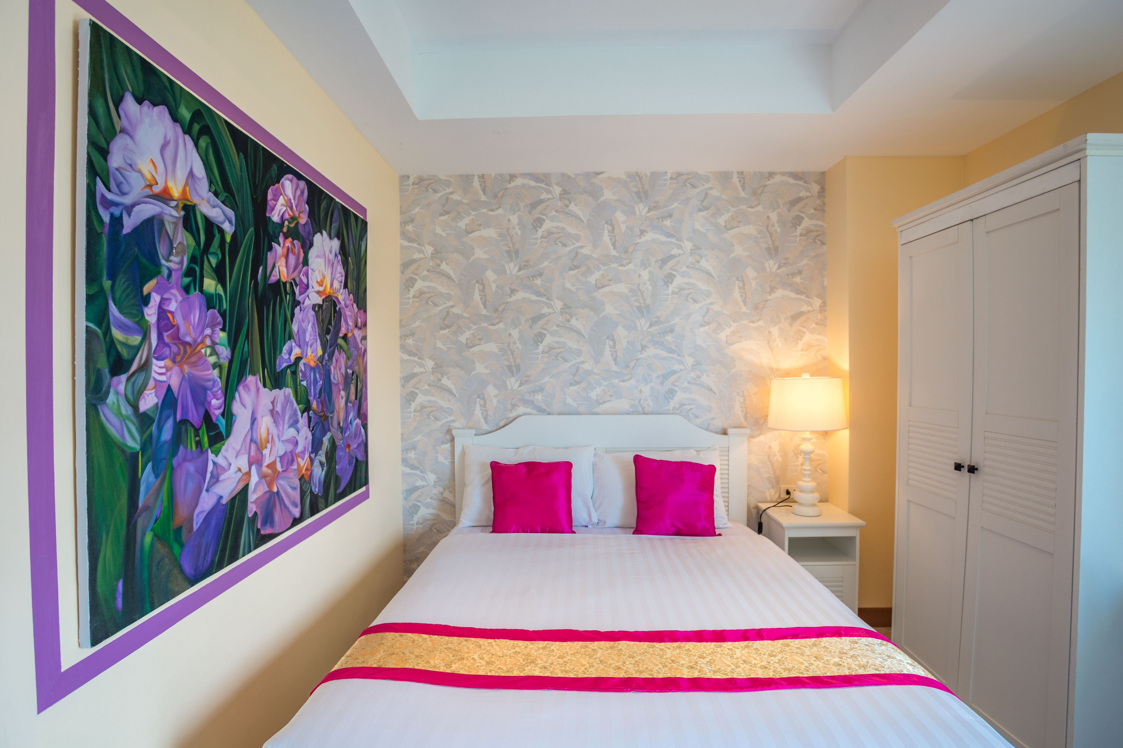 Et hotelværelse med et stort blomsterbillede på væggen, en hvid seng med lyserøde puder og et natbord med en lampe.