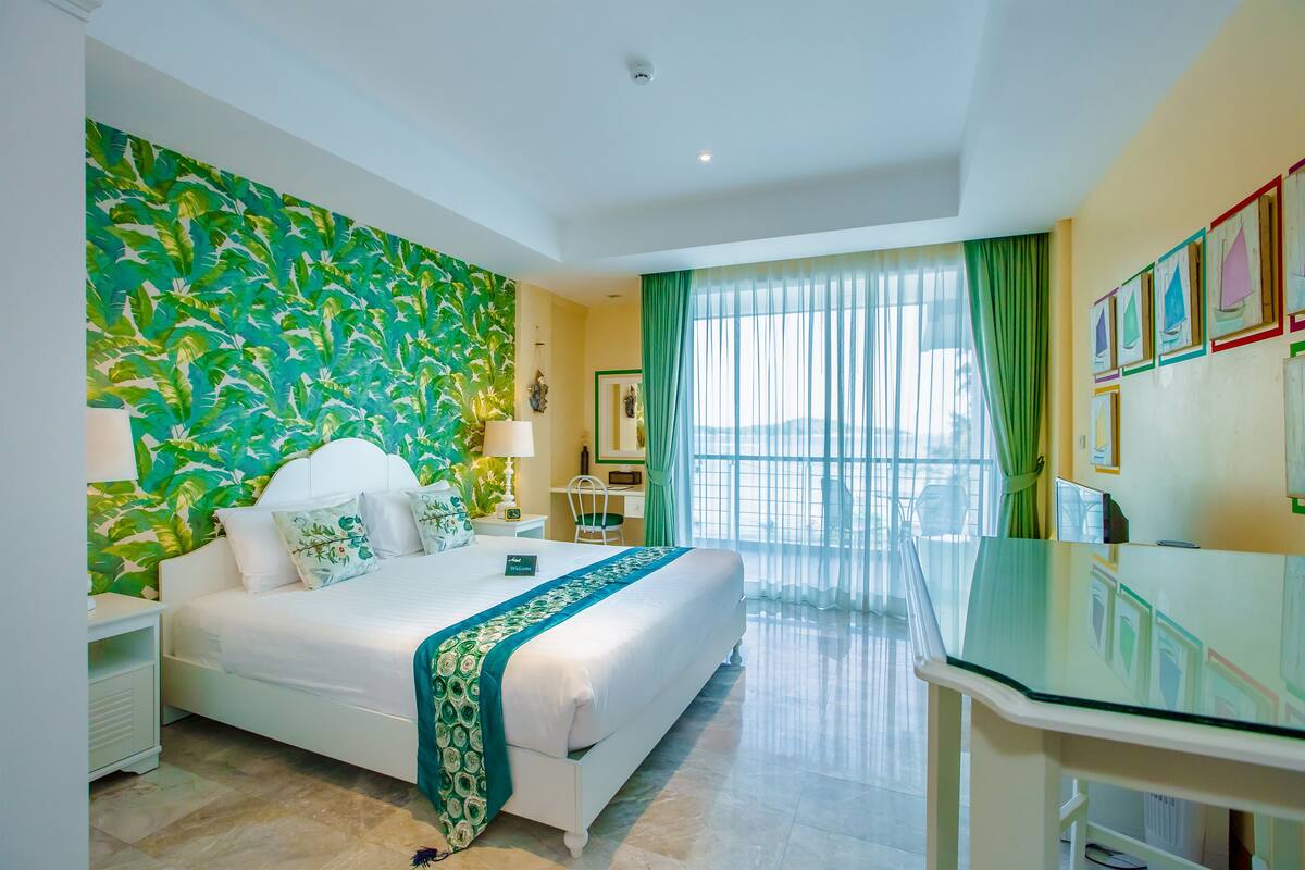 Une chambre d’hôtel avec un grand lit, un bureau avec un plateau en verre, un mur à motifs verts et une vue sur l’extérieur grâce à de grandes fenêtres.