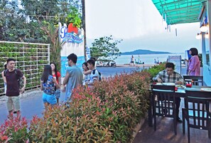A restaurant with outdoor seating overlooking a body of water, a signboard, and a view of the sea. - Almali Rawai Beach Residence (Rawai)