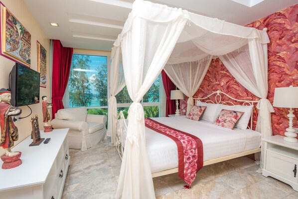 A bedroom with a four-poster canopy bed, a television, a desk with a statue, and a view of trees through the window. - Almali Rawai Beach Residence (Rawai)