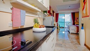 A modern kitchen with a fruit bowl on the counter, a white refrigerator, and a dining area with a white chair and a bed with a red curtain. - Almali Rawai Beach Residence (Rawai)