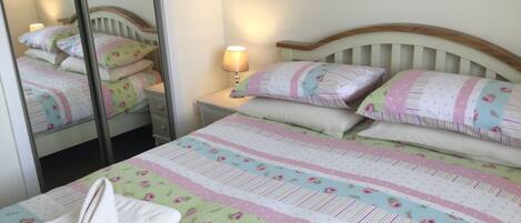 A neatly made bed with floral bedding, a bedside lamp, and a mirror reflecting the room.