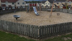 Ein Spielplatz mit Rutsche und Schaukeln, umzäunt mit einem Holzzaun, mit Häusern im Hintergrund.