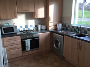 A kitchen with wooden cabinets, a microwave, a stove, a washing machine, and a window with a view of the outside. - Inverness Apartments - Morning Field (Inverness)