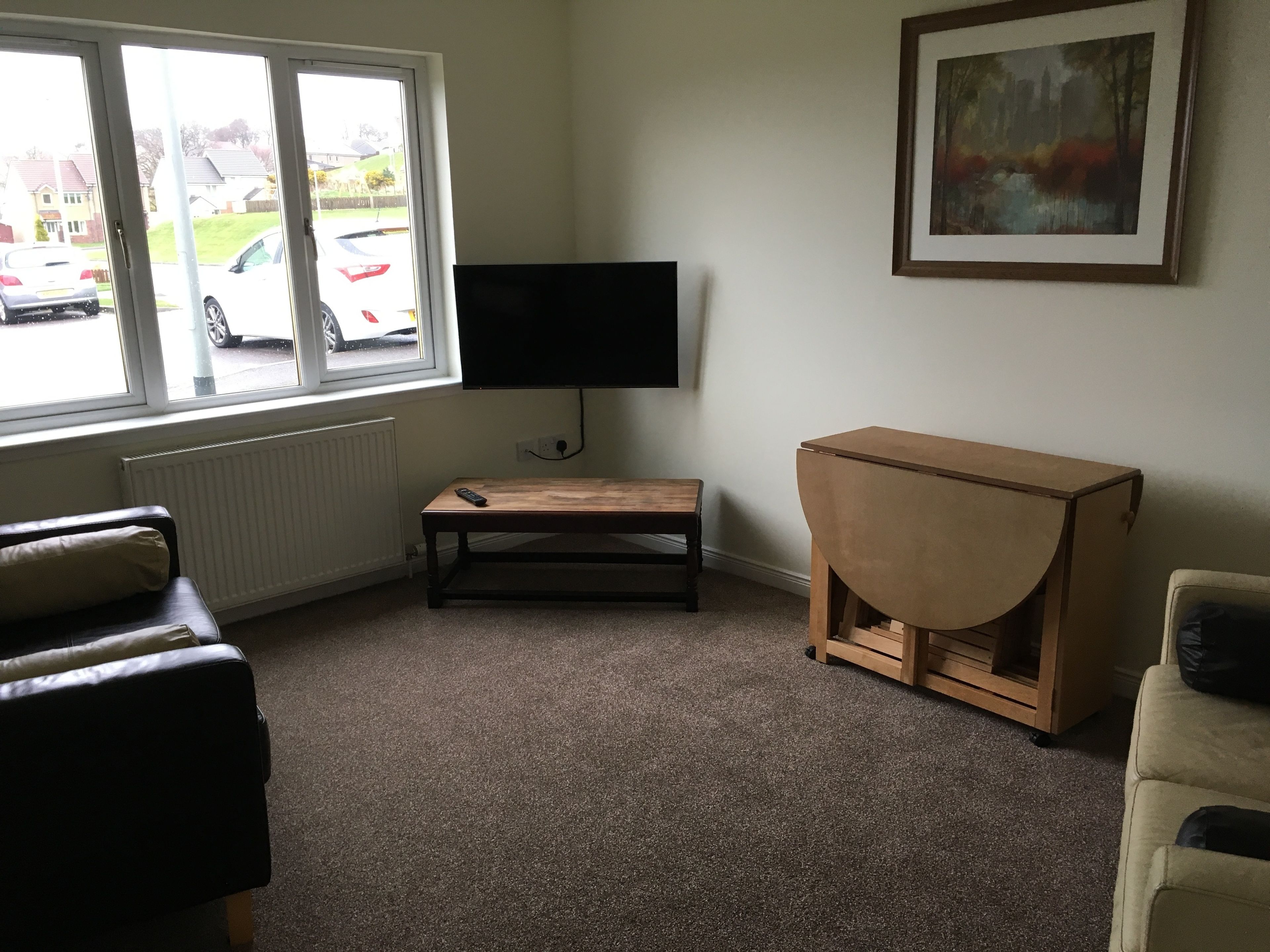 A living room with a TV on a stand, a wooden table, a sofa, and a window with a view of parked cars.