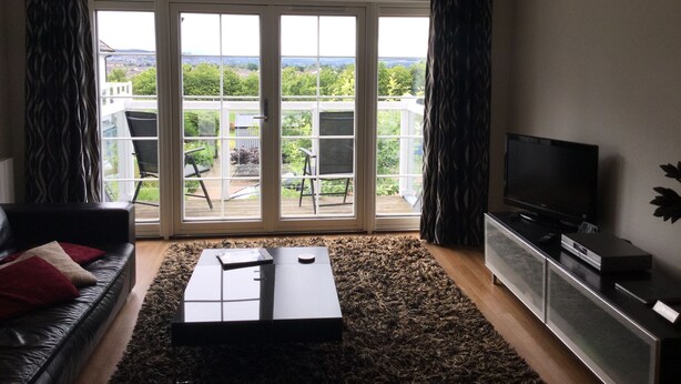 A living room with a sofa, coffee table, TV, and a sliding glass door leading to a balcony with outdoor furniture. - Inverness Apartments - Castlefield (Inverness)