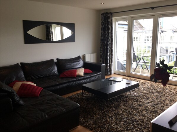 A living room with a black leather sofa, a coffee table, and a large window with curtains. - Inverness Apartments - Castlefield (Inverness)
