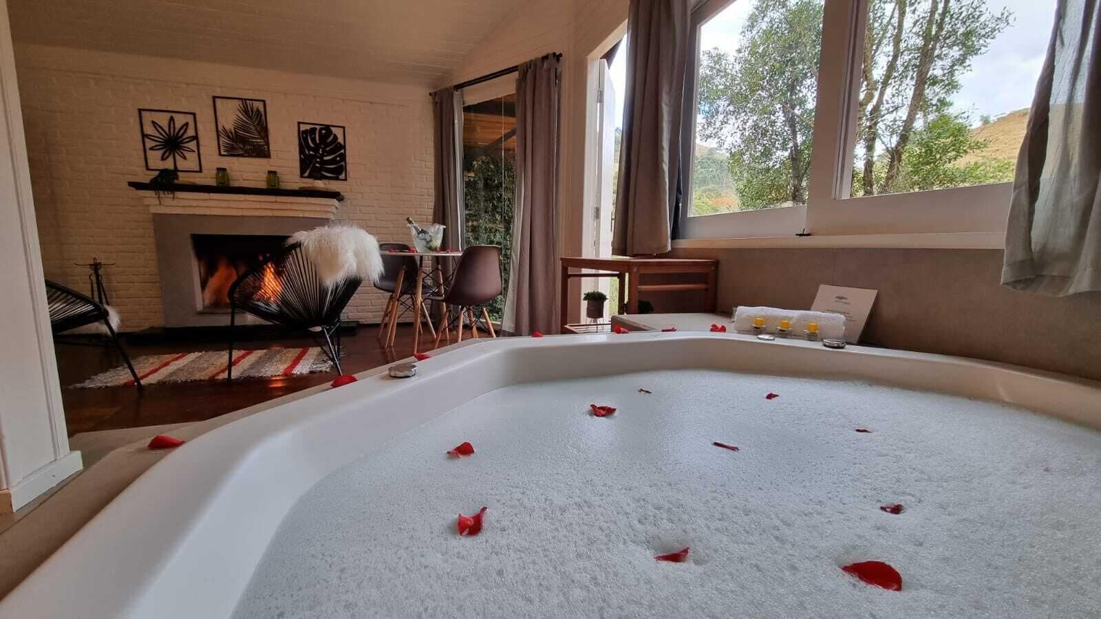 A room with a large bathtub filled with water and rose petals, a fireplace, and a view of trees through large windows.