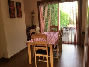 En matplats med ett bord dukat för fyra personer, trästolar, en vas med blommor och en utsikt genom ett fönster med persienner.
