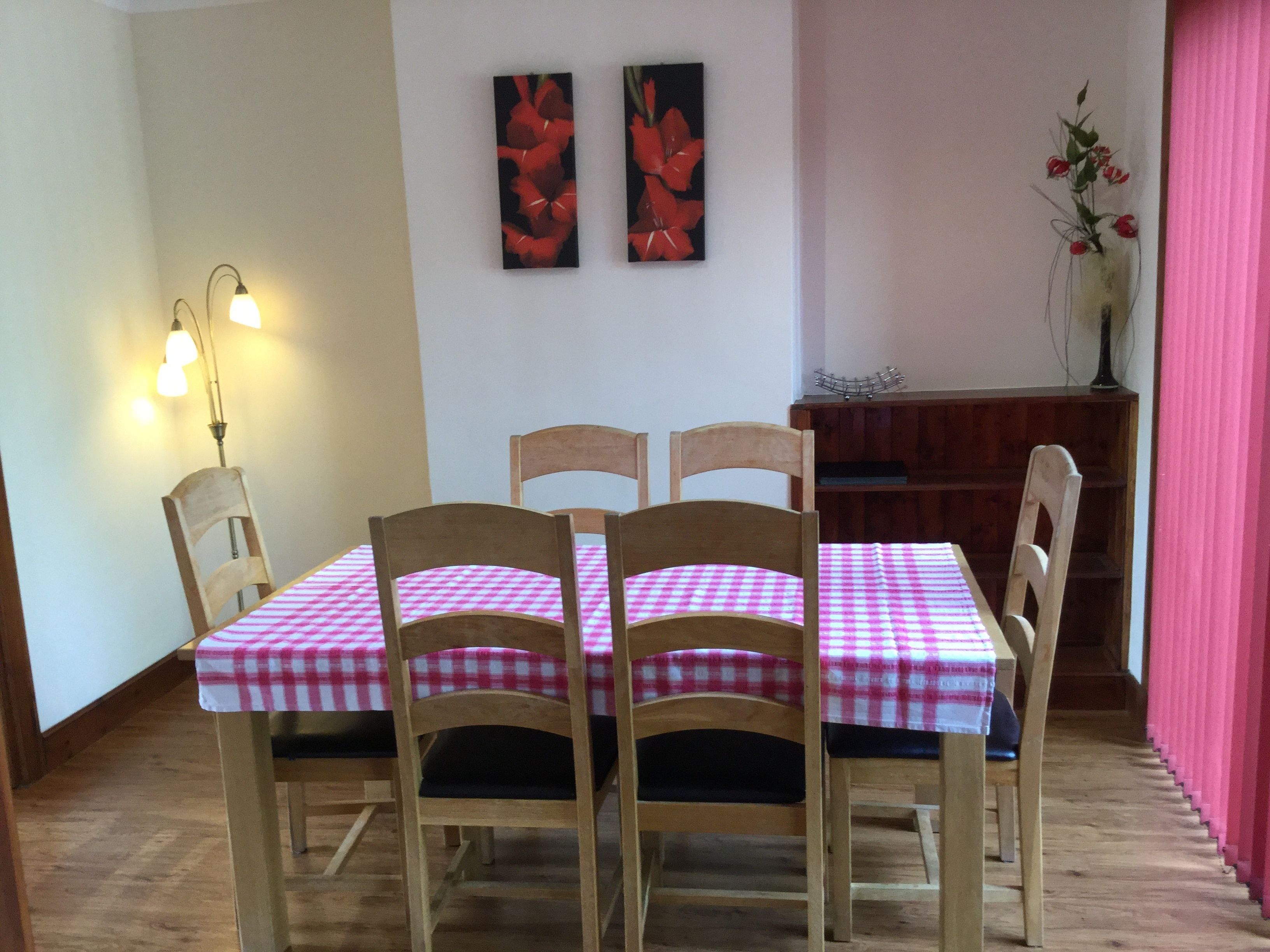 A dining area with a table set for four, wooden chairs, a checkered tablecloth, two wall-mounted paintings, a lamp, and a vase with flowers.