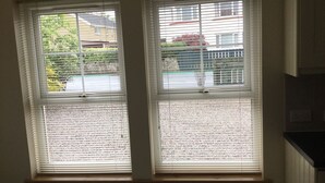 Two windows with blinds, a radiator below, and a view of a residential area with houses and greenery. - Inverness Apartments - New Cottage (Inverness)