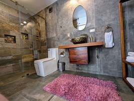 Un baño moderno con ducha de vidrio, inodoro blanco, tocador de madera con espejo y alfombra rosa en el suelo.