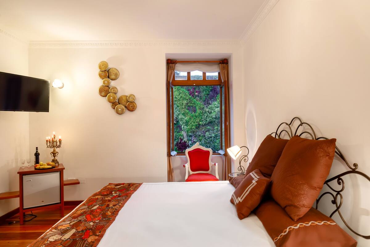 Hotelkamer met een bed, een televisie, een tafeltje met een kandelaar, een raam met uitzicht op groen, en een rode stoel.