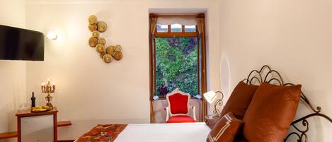 Habitación de hotel con cama, televisor, una mesita con un candelabro, ventana con vistas a la vegetación y una silla roja.