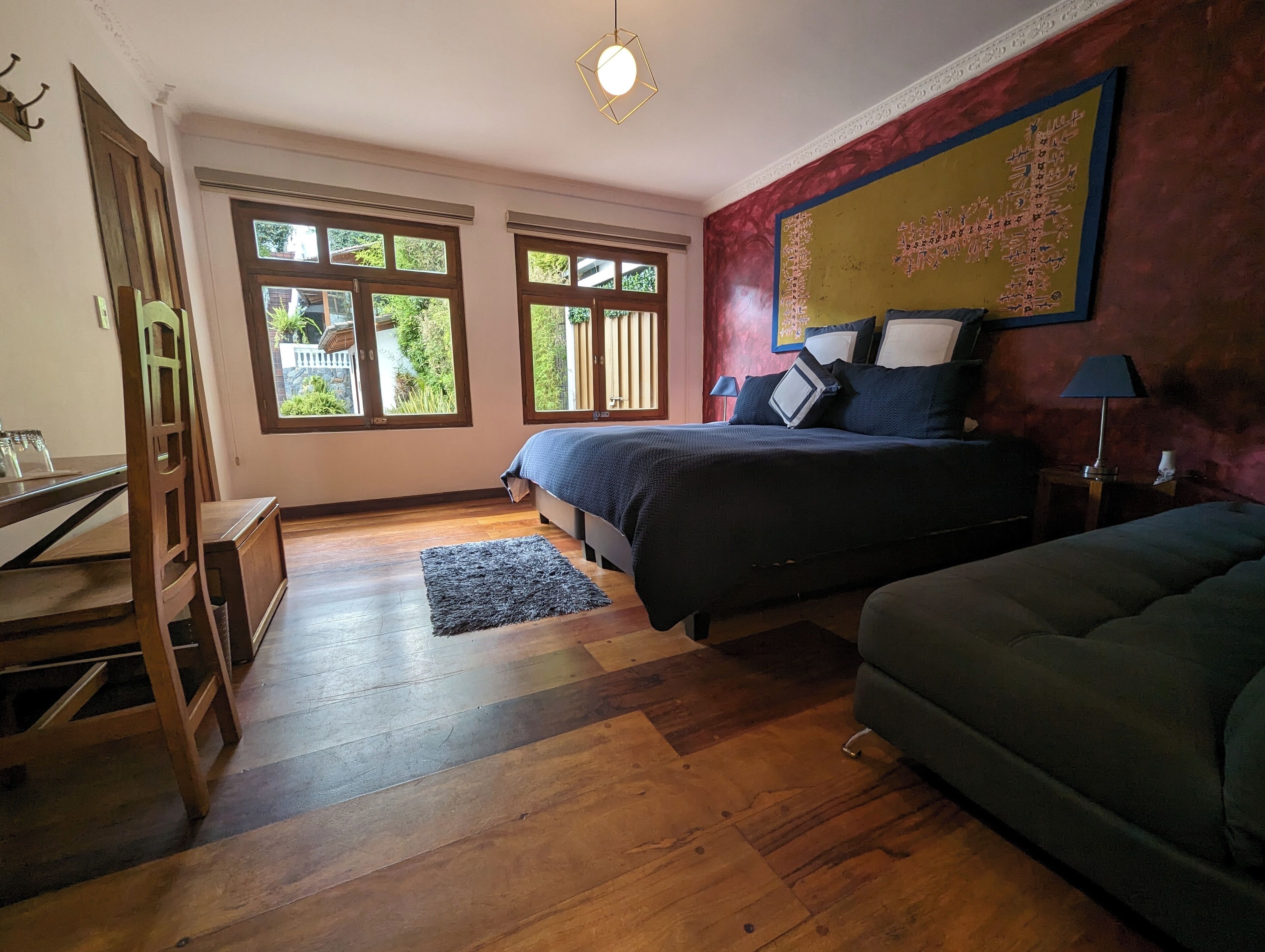 A bedroom with a bed, a desk, a chair, and a sofa. There are windows and a view of greenery outside.