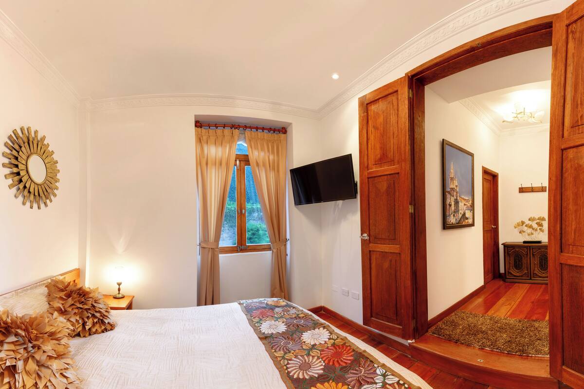 Een slaapkamer met een bed, een televisie, een raam met gordijnen en een houten deur.