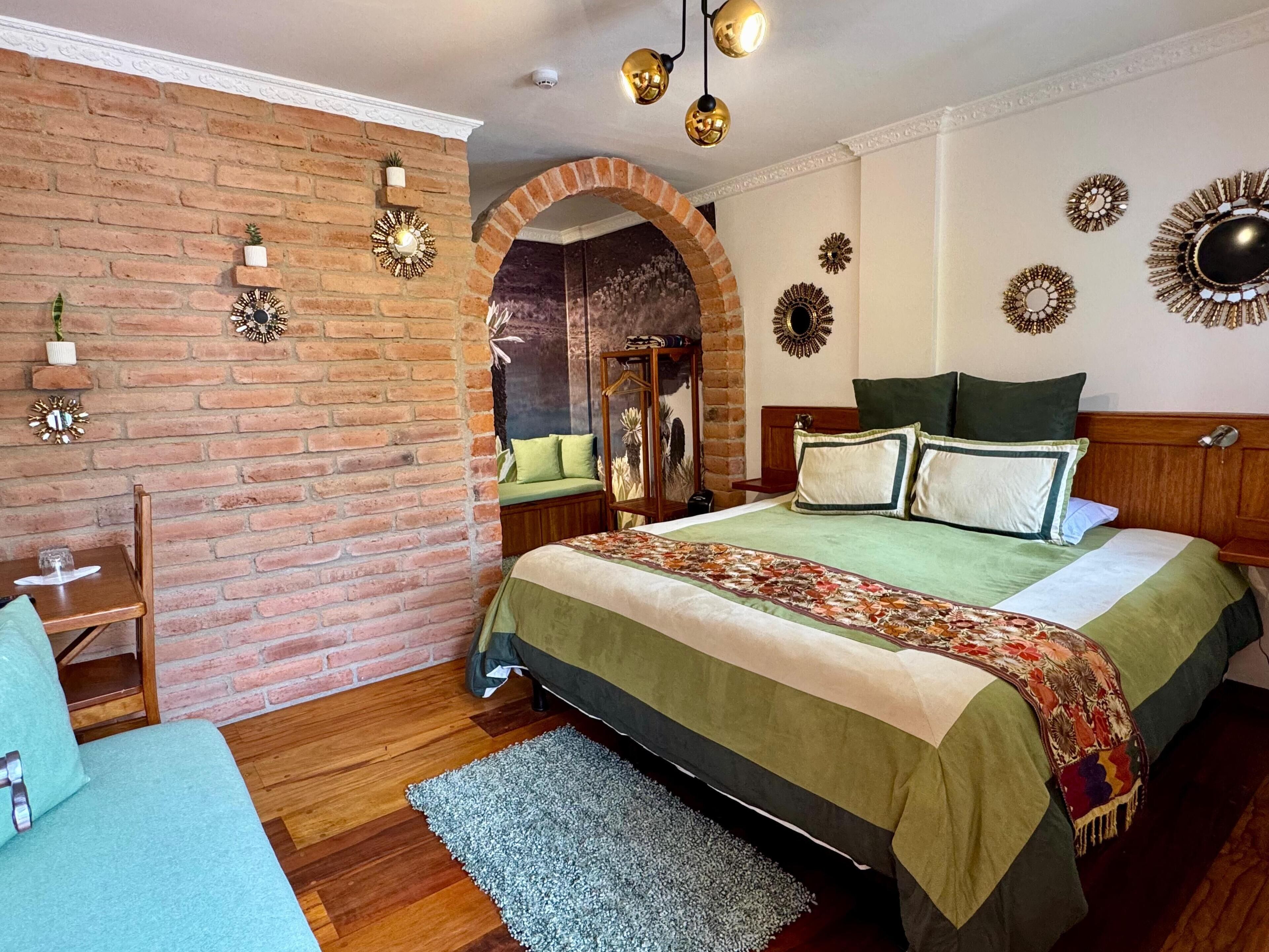 A bedroom with a brick wall, a wooden bed, a green bedspread, and decorative wall mirrors.