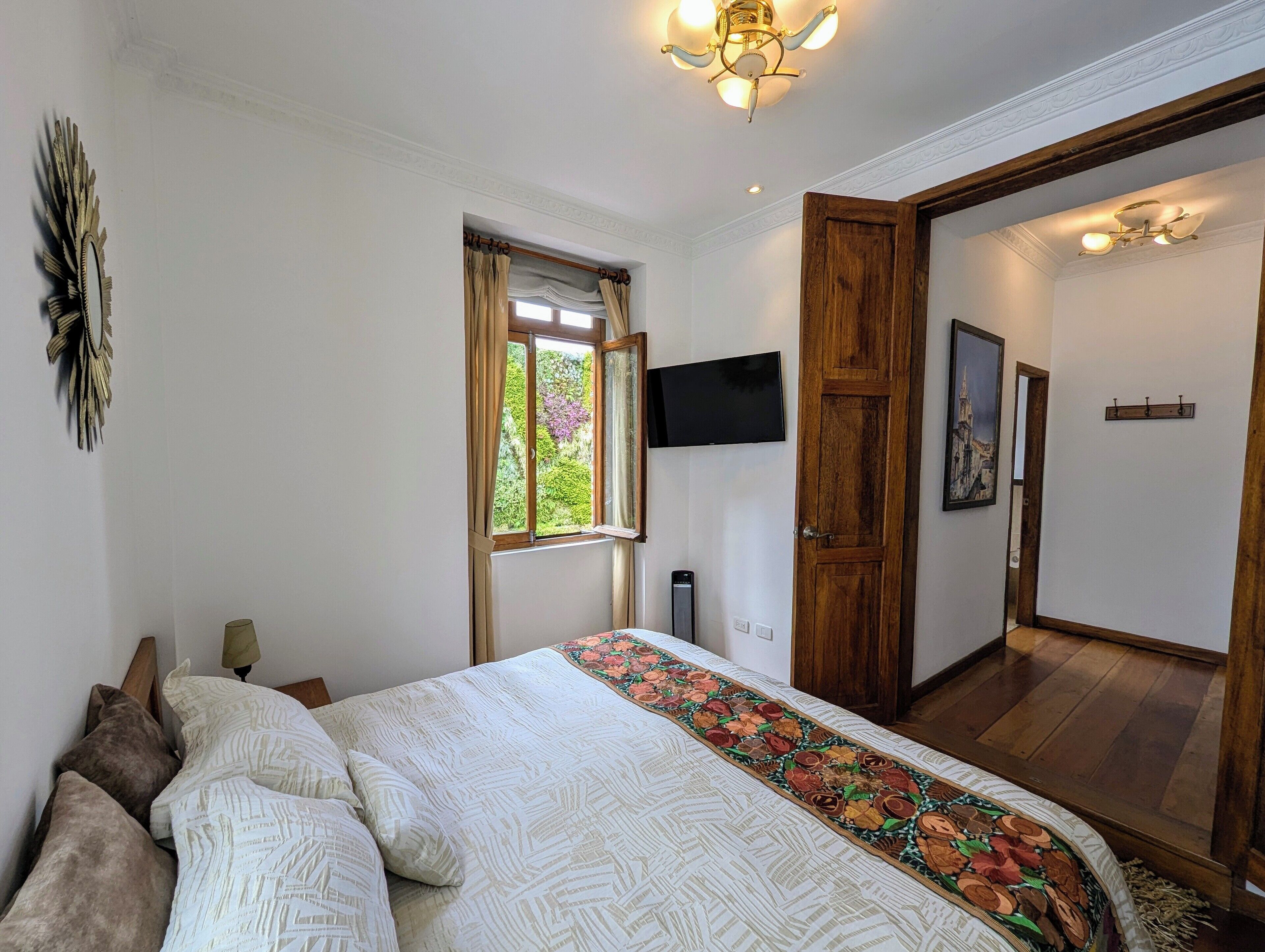 Un dormitorio con cama, televisor montado en la pared, ventana con cortinas y puerta que da a otra habitación.