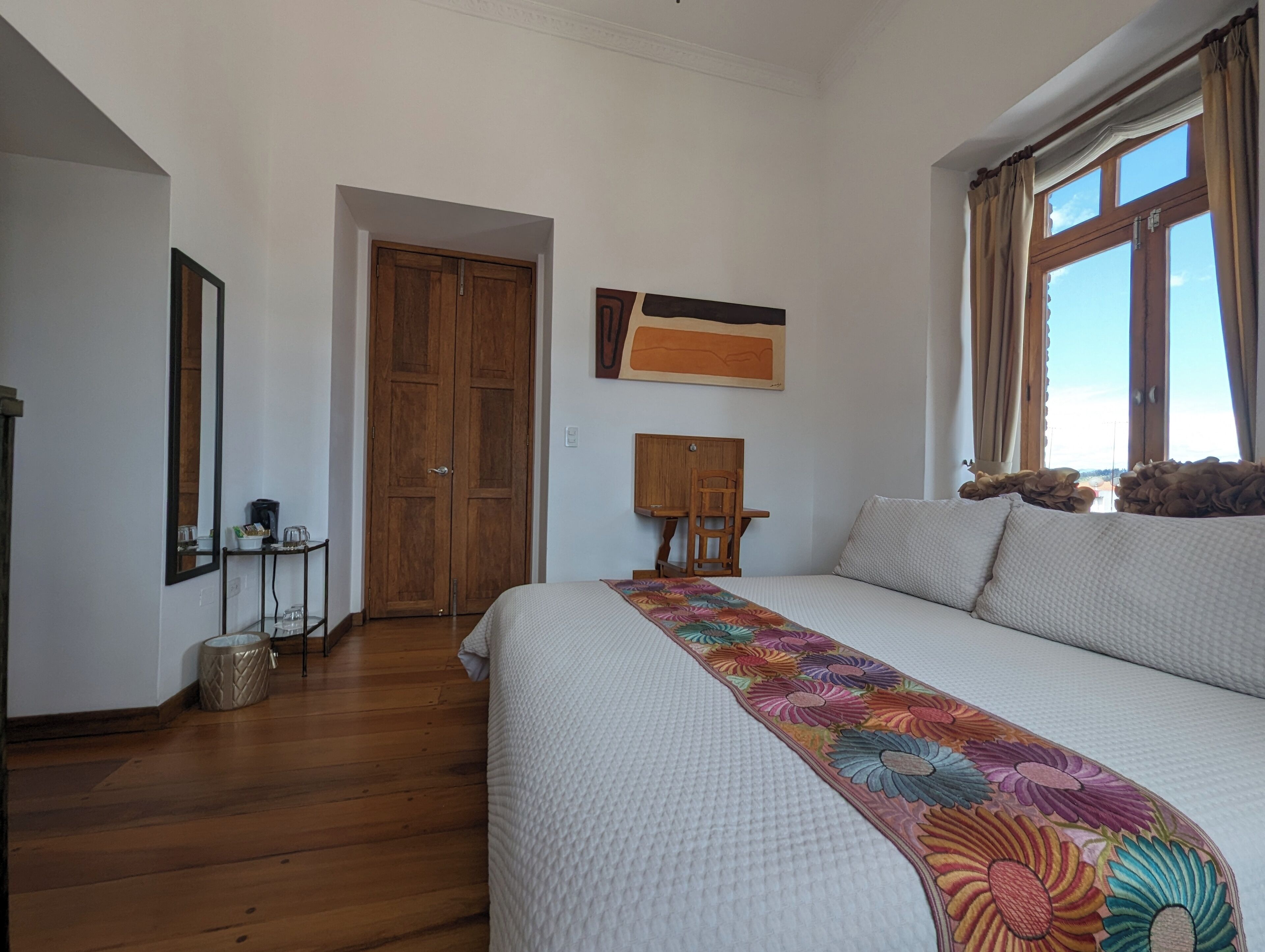 A bedroom with a large bed, a wooden wardrobe, a small desk, a chair, and a window with curtains.
