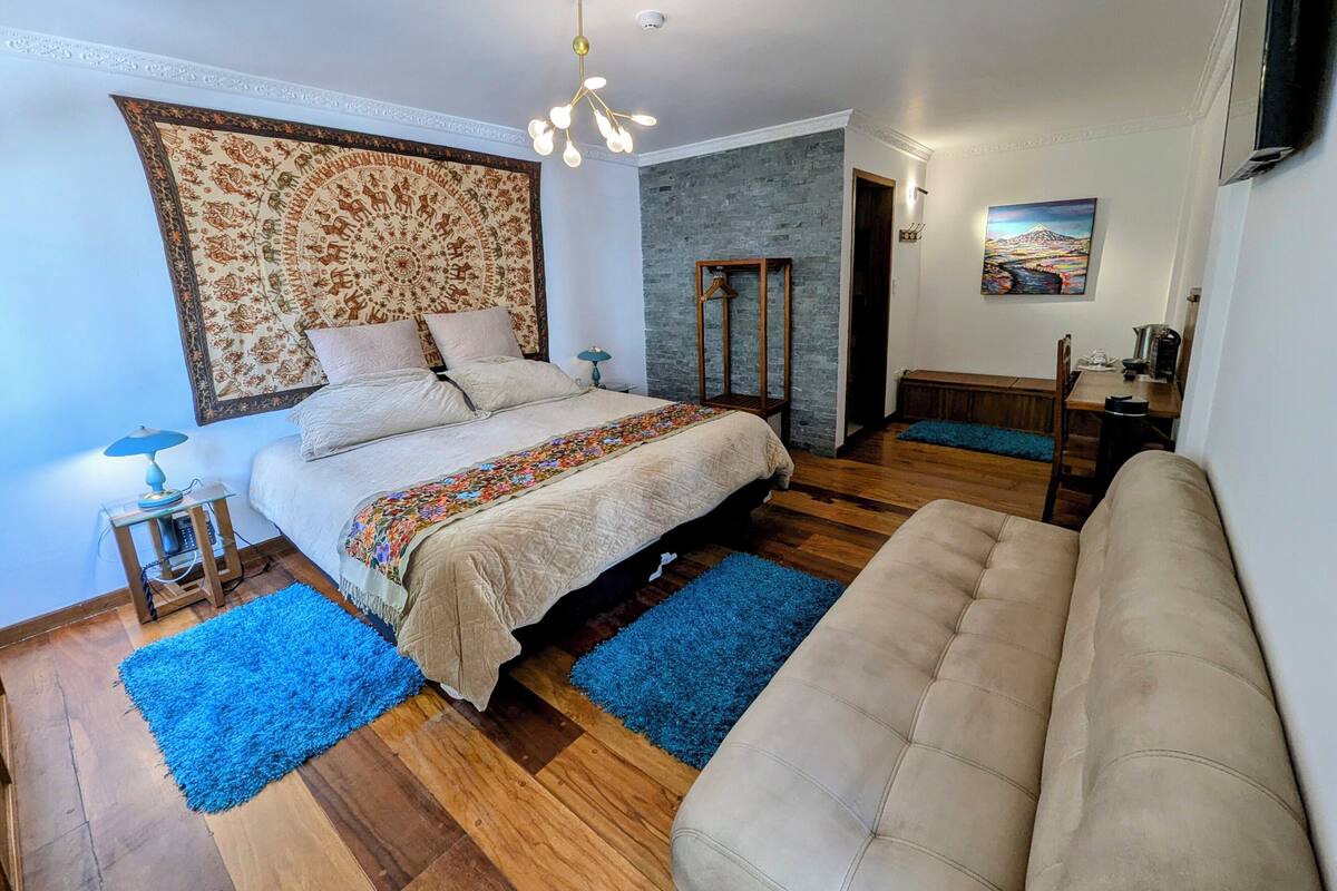 Een slaapkamer met een bed, een bank, een blauw vloerkleed, een houten vloer, een gestreept hoofdeinde, een hanglamp en een schilderij aan de muur.