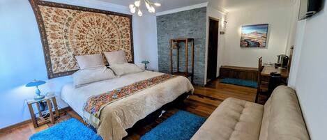 Un dormitorio con cama, sofá, alfombra azul, suelo de madera, cabecero estampado, lámpara colgante y un cuadro en la pared.