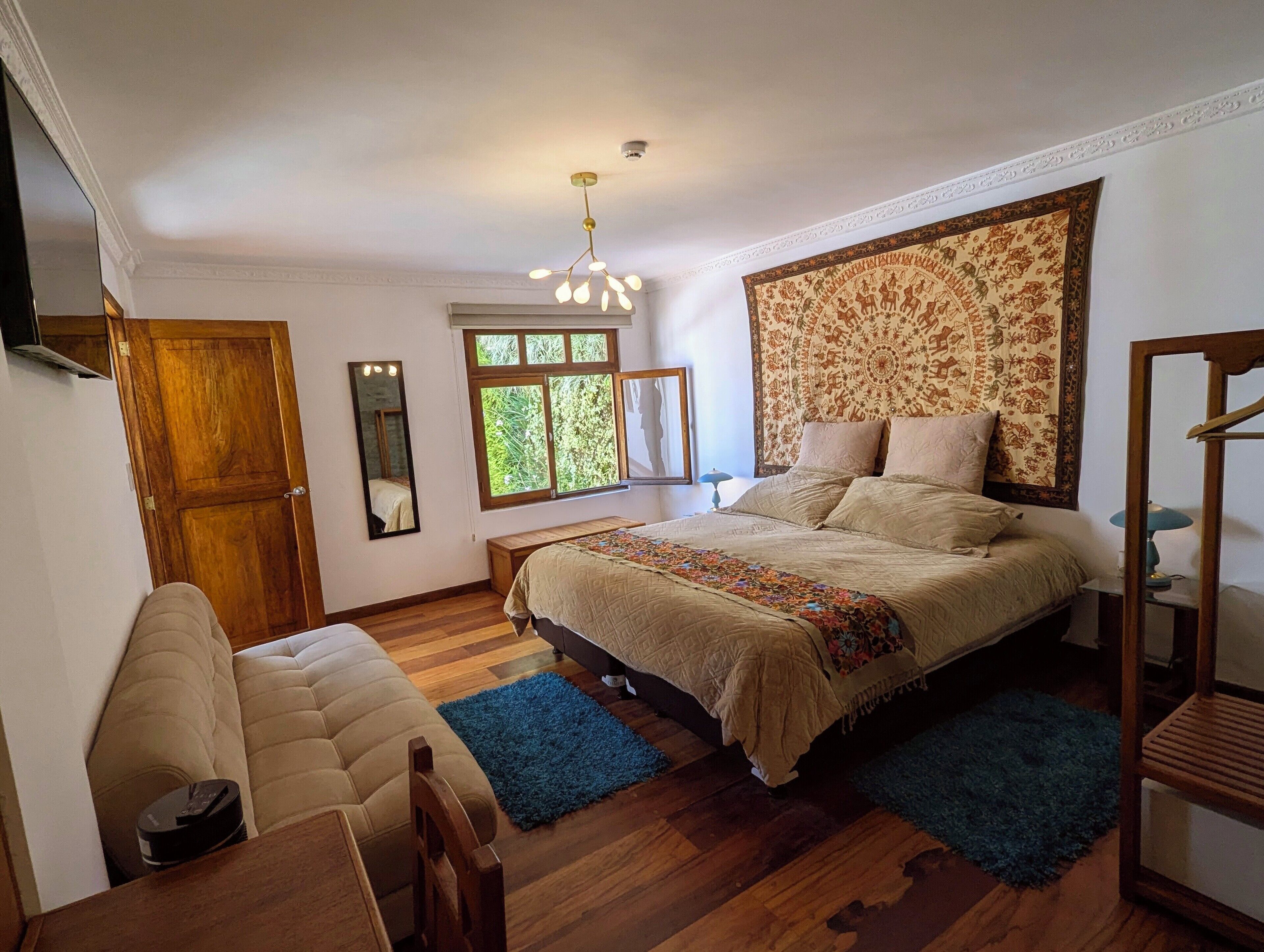 A bedroom with a large bed, a wooden wardrobe, a small table, and a chair. There is a window with a view of greenery outside.