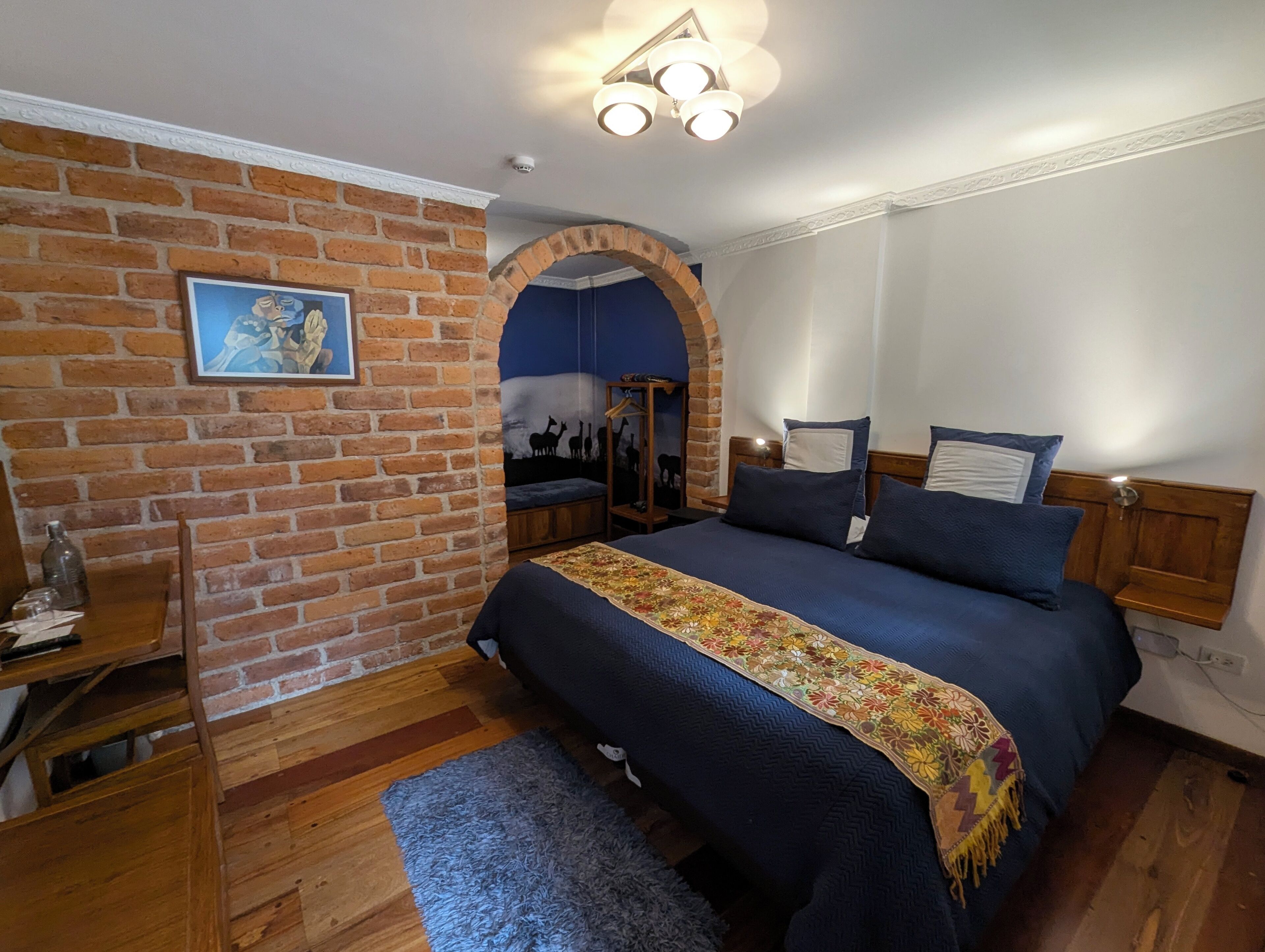 A bedroom with a brick wall, a bed with a floral bedspread, a wooden headboard, a nightstand, and a wall-mounted shelf.