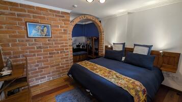 A bedroom with a brick wall, a bed with a floral bedspread, a wooden headboard, a nightstand, and a wall-mounted shelf.