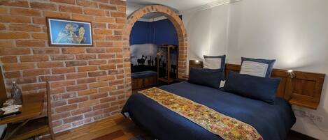 A bedroom with a brick wall, a bed with a floral bedspread, a wooden headboard, a nightstand, and a wall-mounted shelf.