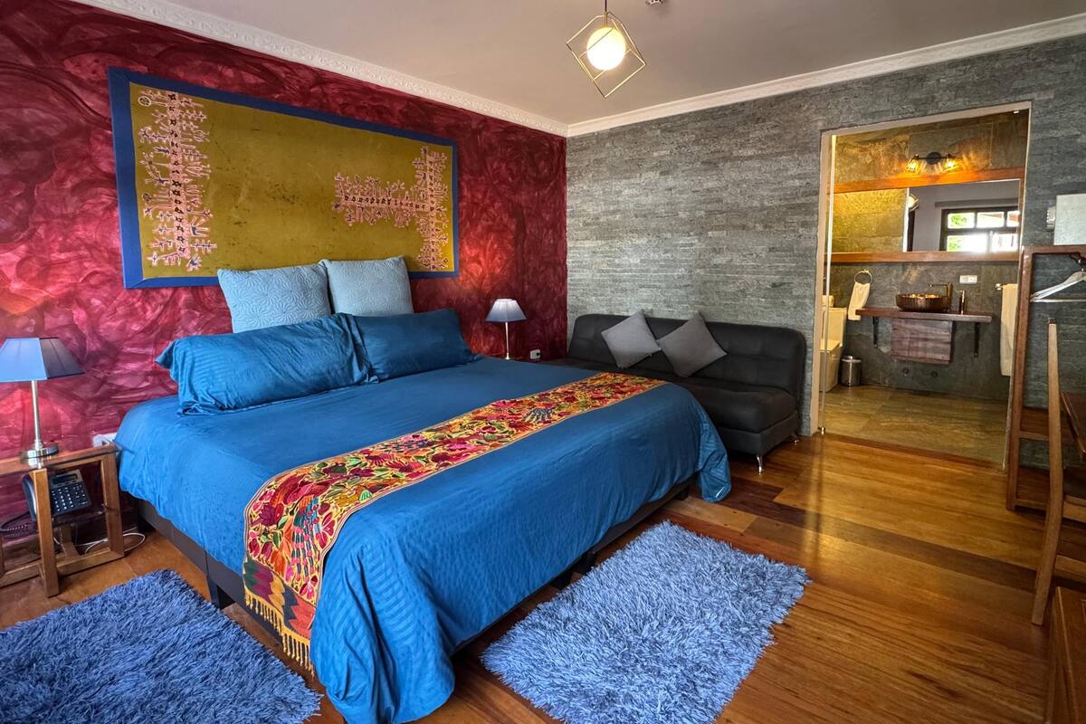 Een moderne slaapkamer met een groot bed, een zwarte bank, een kitchenette en een rode accentmuur.