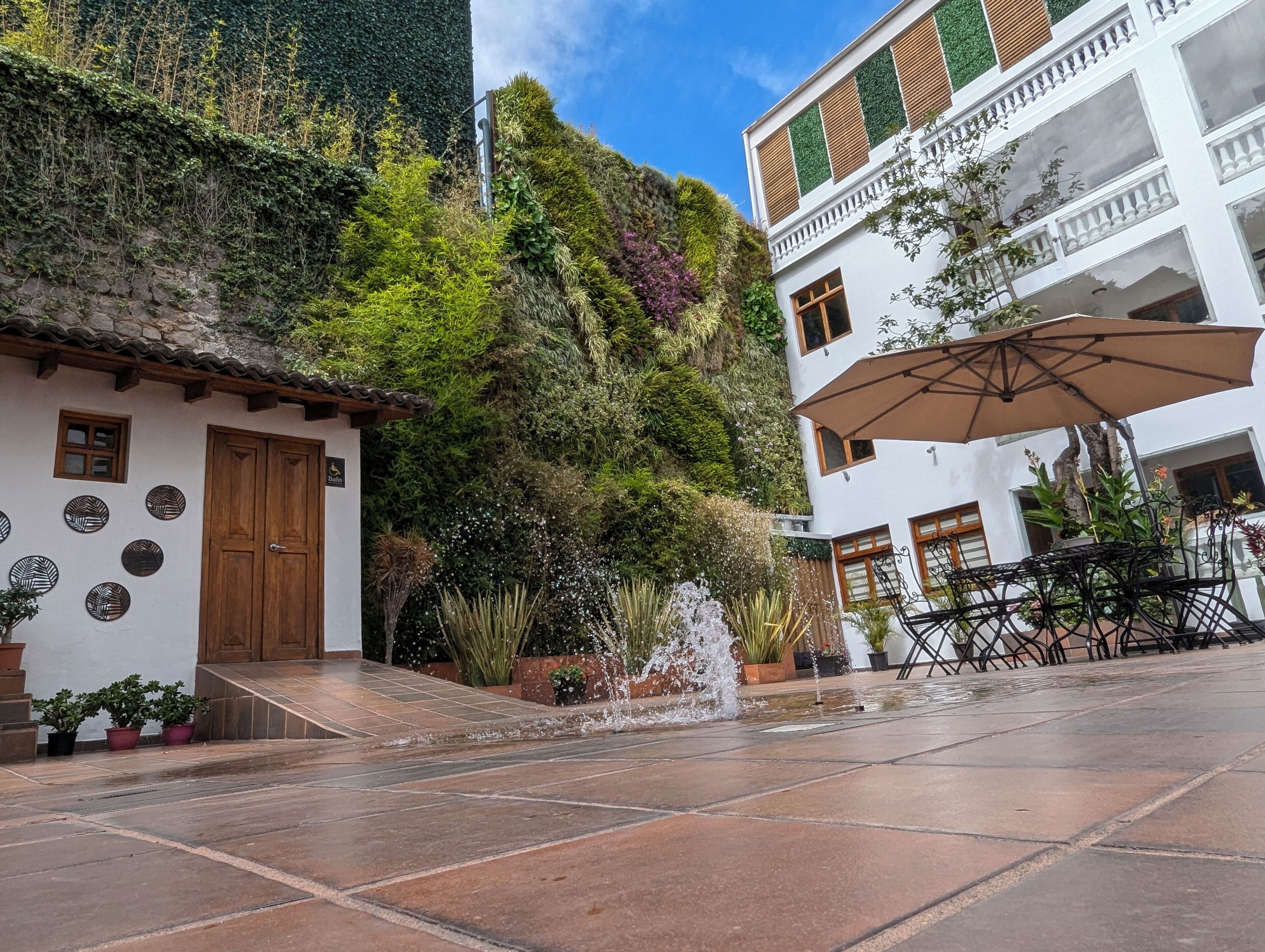 Une cour avec une fontaine, des plantes en pot et un bâtiment doté d’une porte en bois et d’un panneau indiquant qu’il est accessible aux personnes à mobilité réduite.
