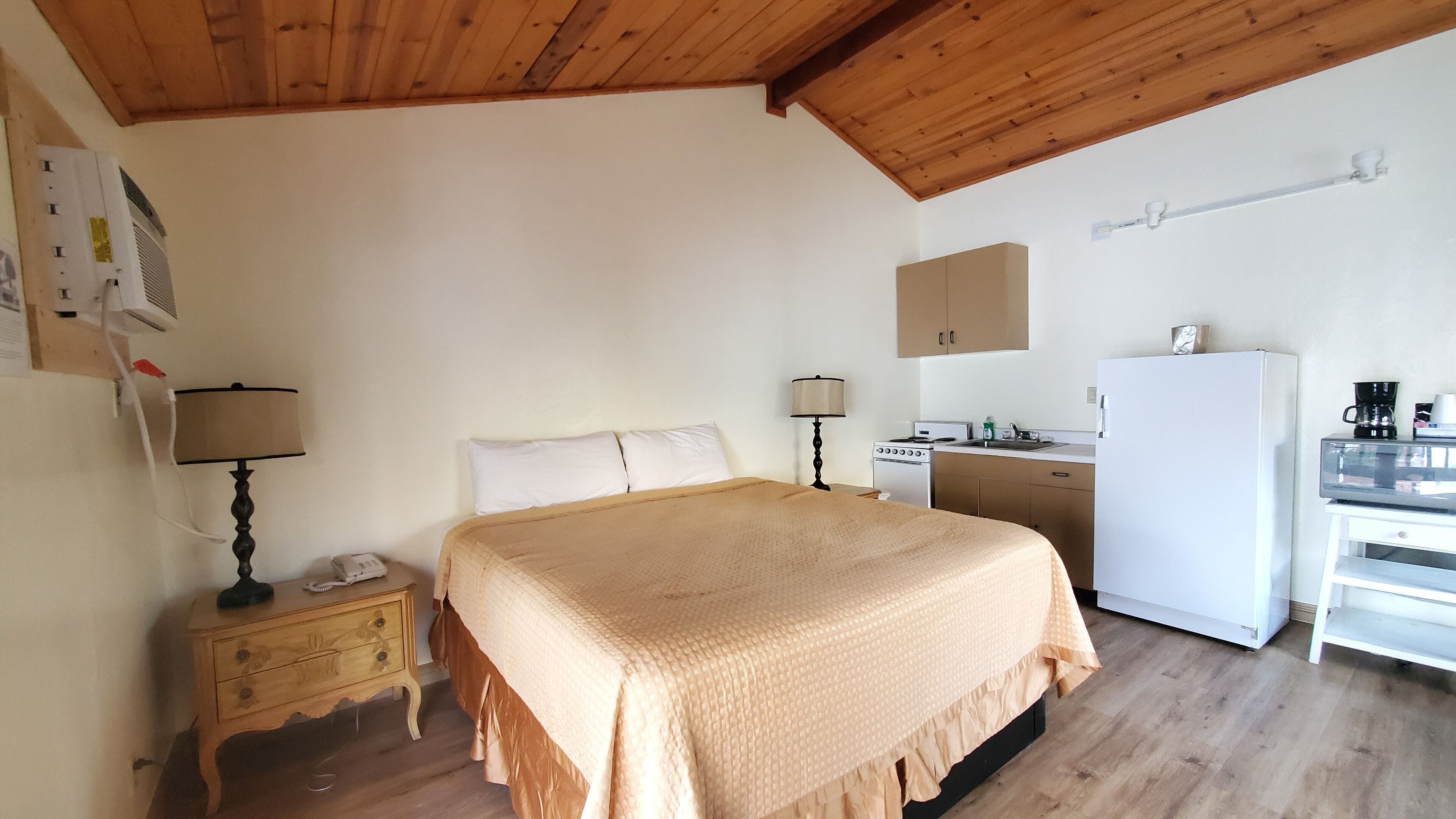 Un dormitorio con cama, mesitas de noche, una lámpara, un pequeño escritorio con silla, kitchenette y refrigerador.