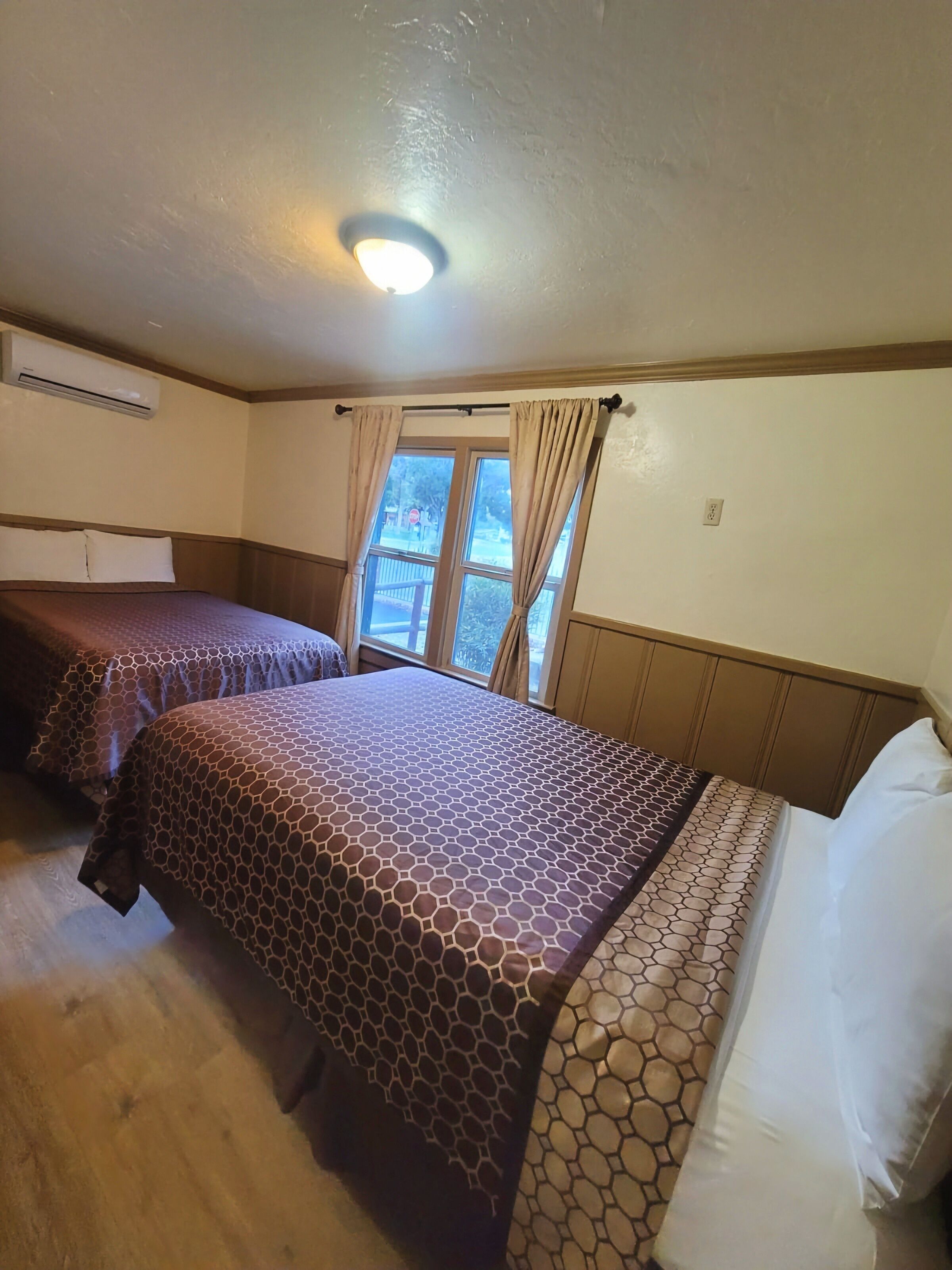 A hotel room with two beds, a window with curtains, an air conditioning unit, and a ceiling light.