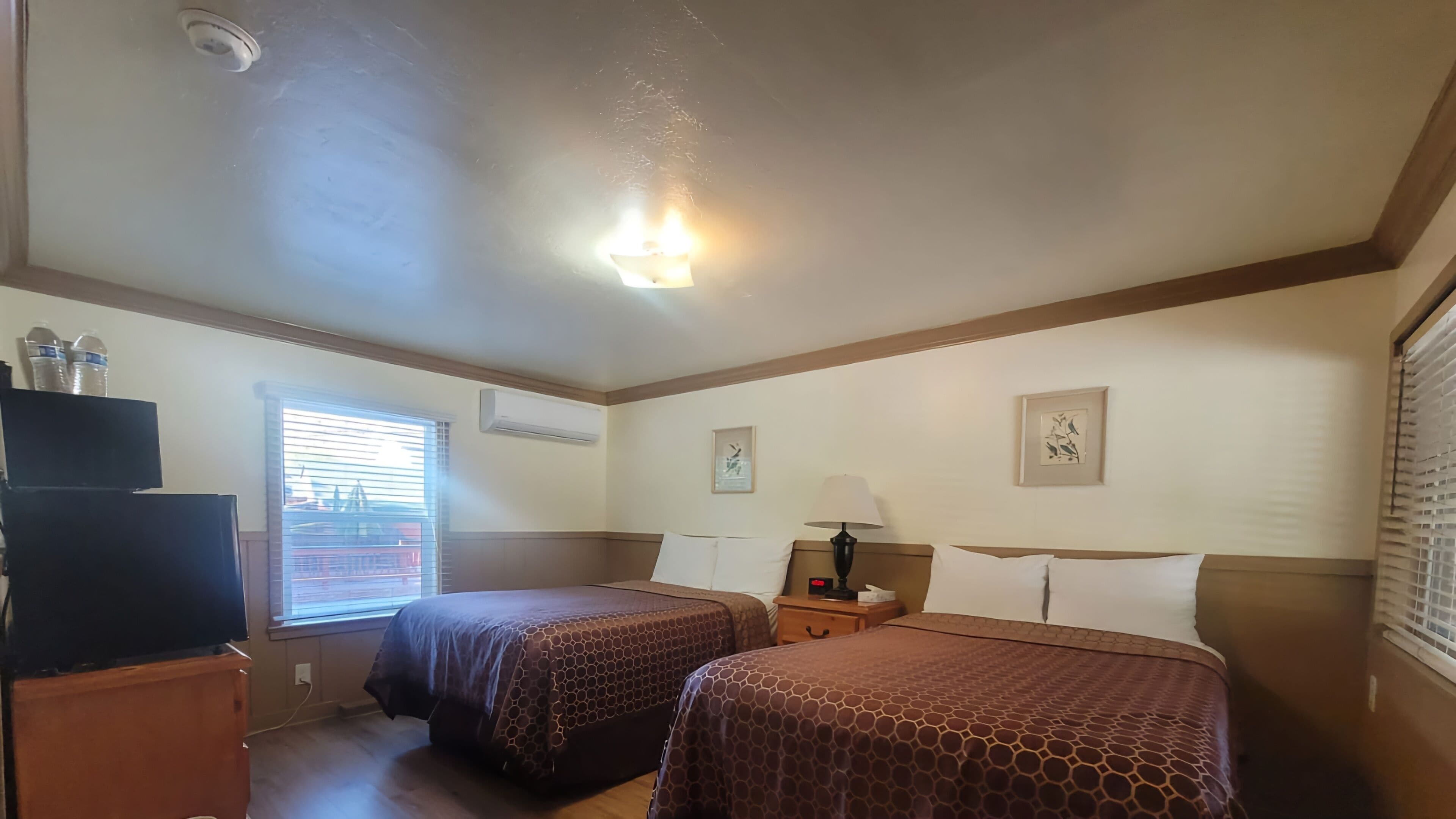 A hotel room with two beds, a TV, a desk, and a window with blinds.