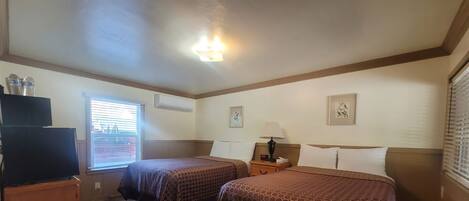 A hotel room with two beds, a TV, a desk, and a window with blinds.