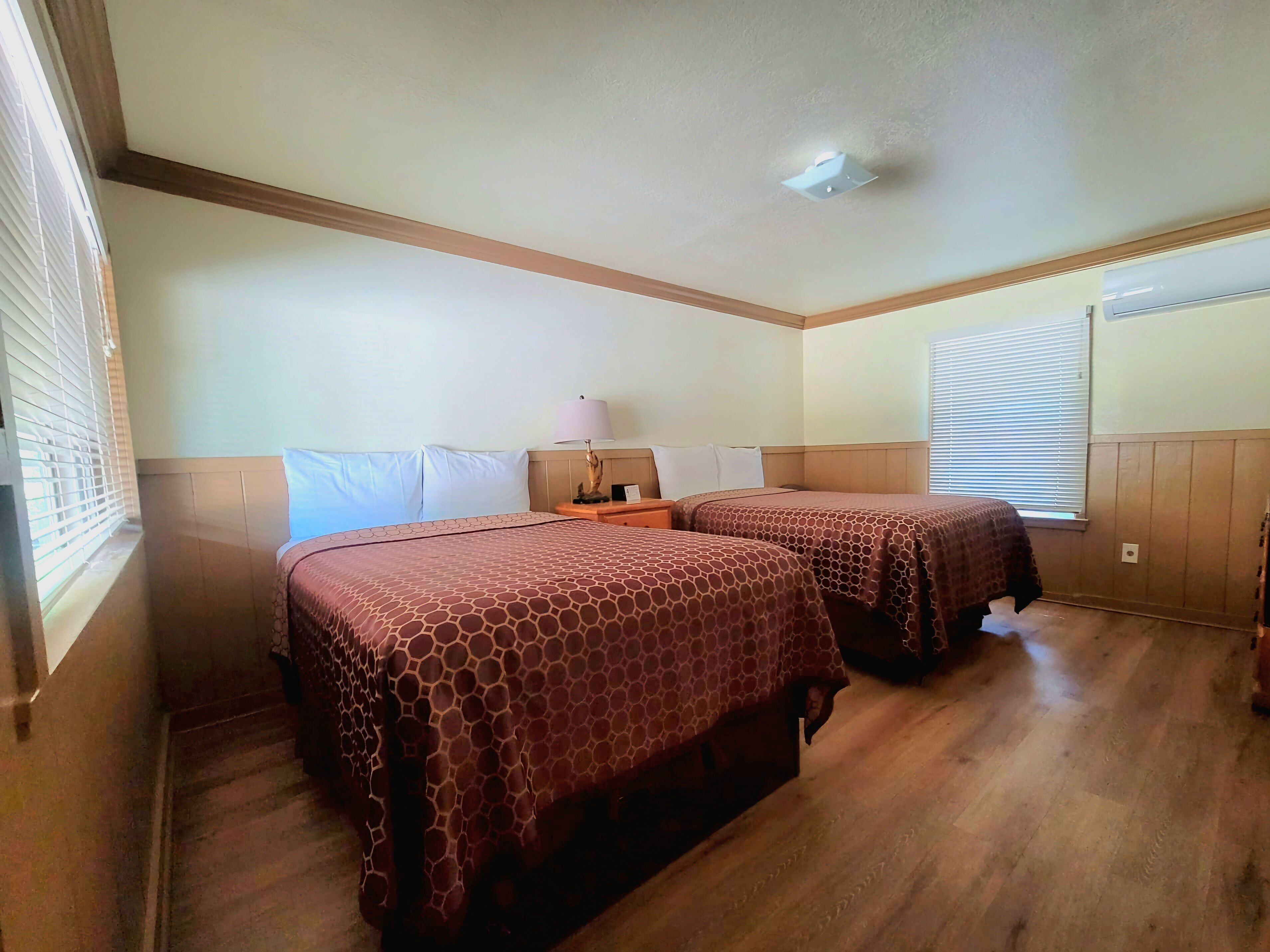A hotel room with two beds, wooden flooring, a ceiling fan, and a window with blinds.