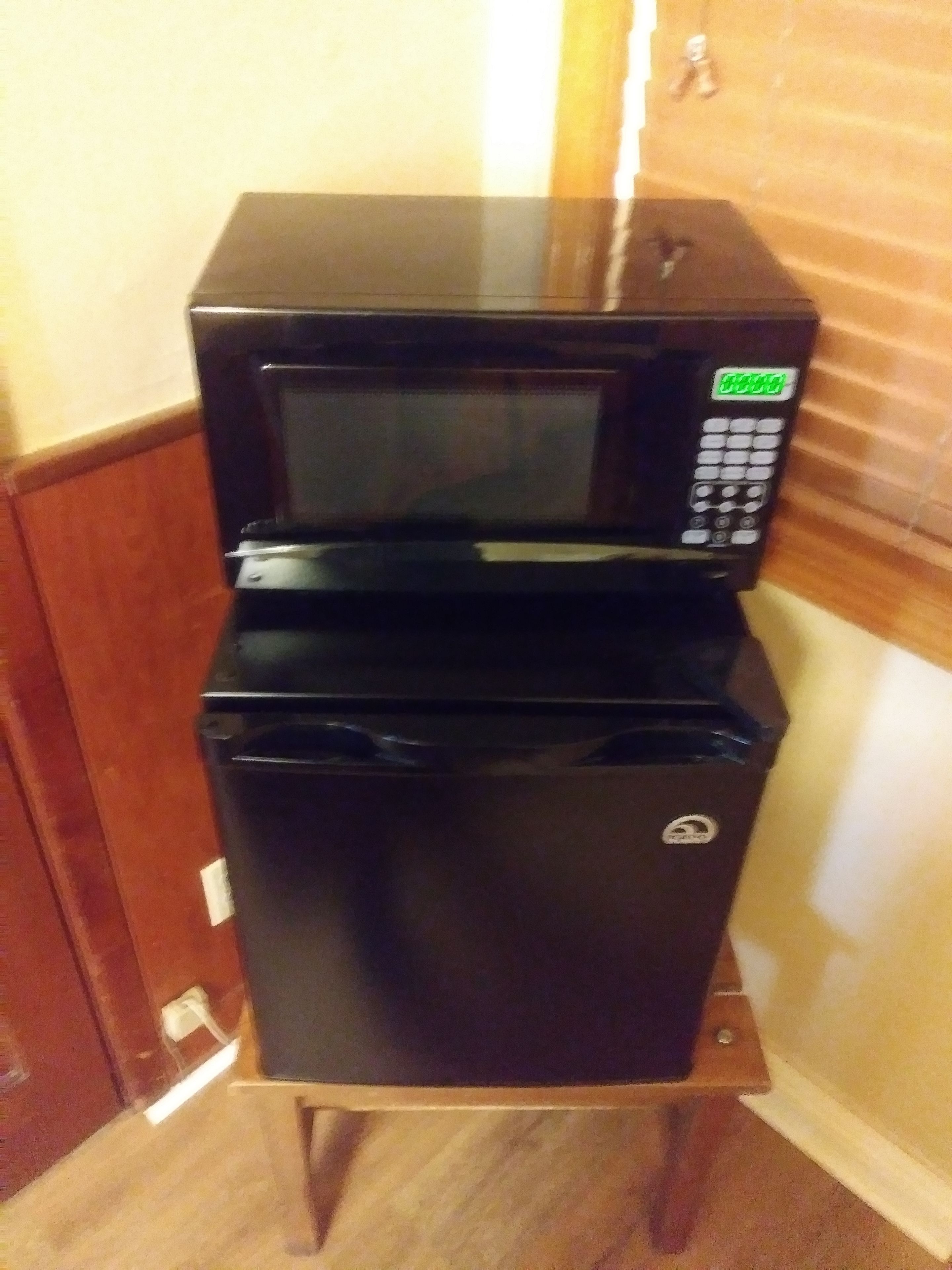 A black microwave and refrigerator stacked on a wooden cabinet in a room with wooden walls and a wooden floor.