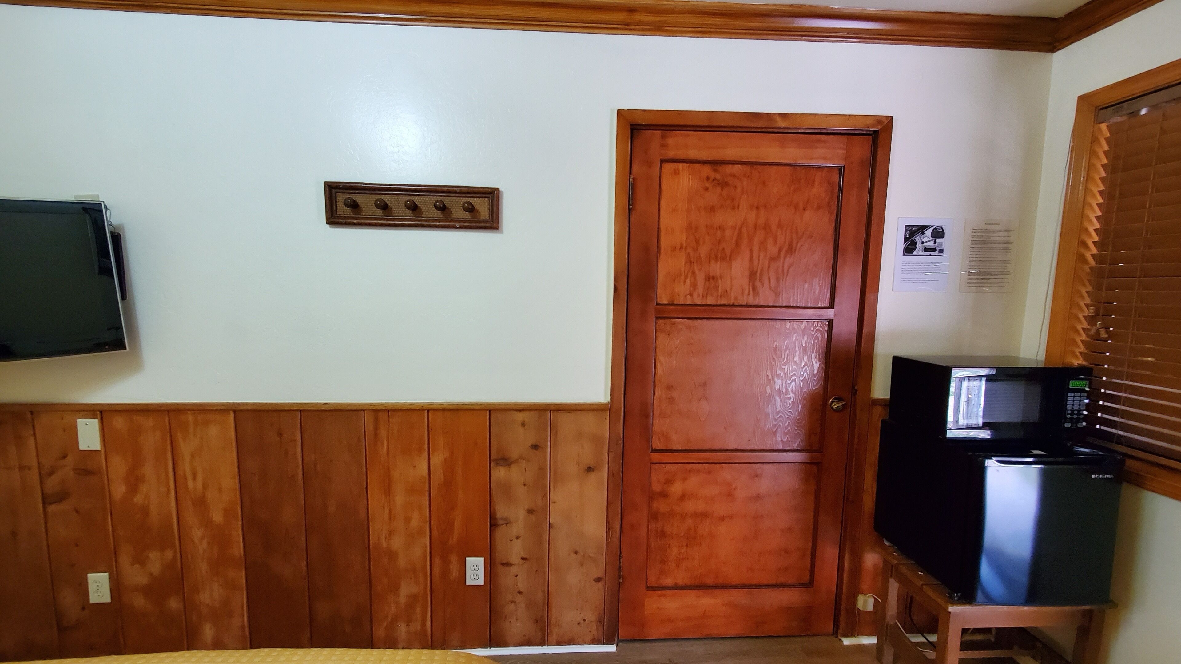 A room with a wooden door, a flat-screen TV mounted on the wall, and a microwave on a wooden stand.