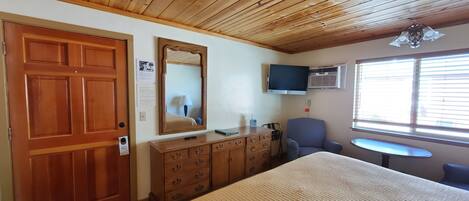 A room with a wooden ceiling, a large mirror, a television, a desk, a chair, and a window with blinds.