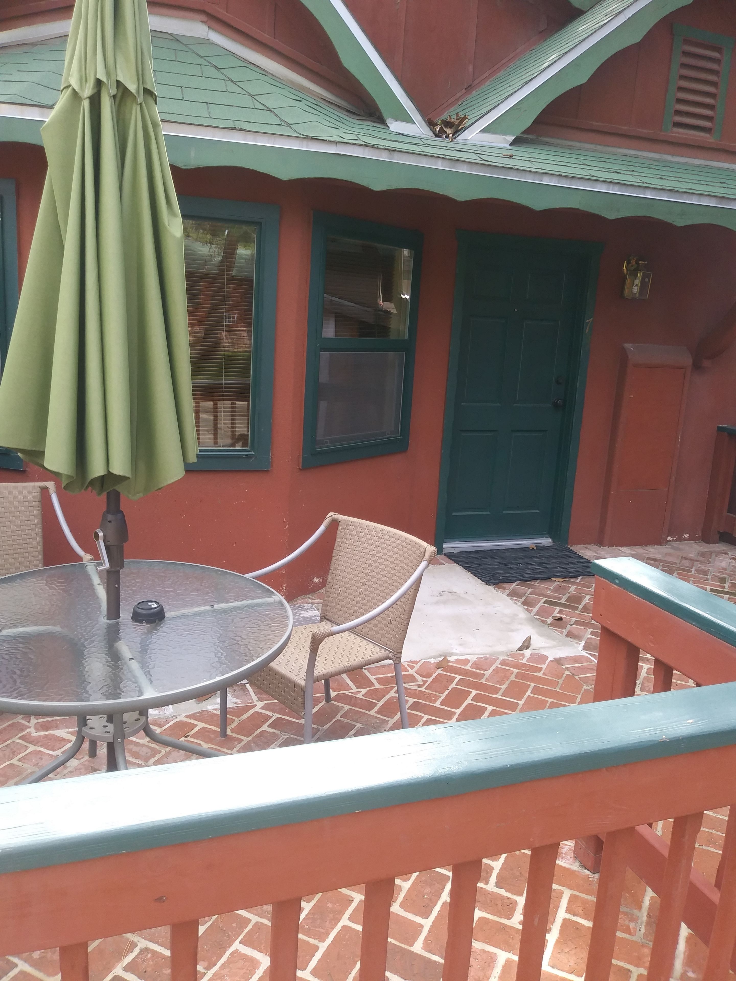 Un patio con una mesa redonda de vidrio, dos sillas de mimbre y una sombrilla verde.