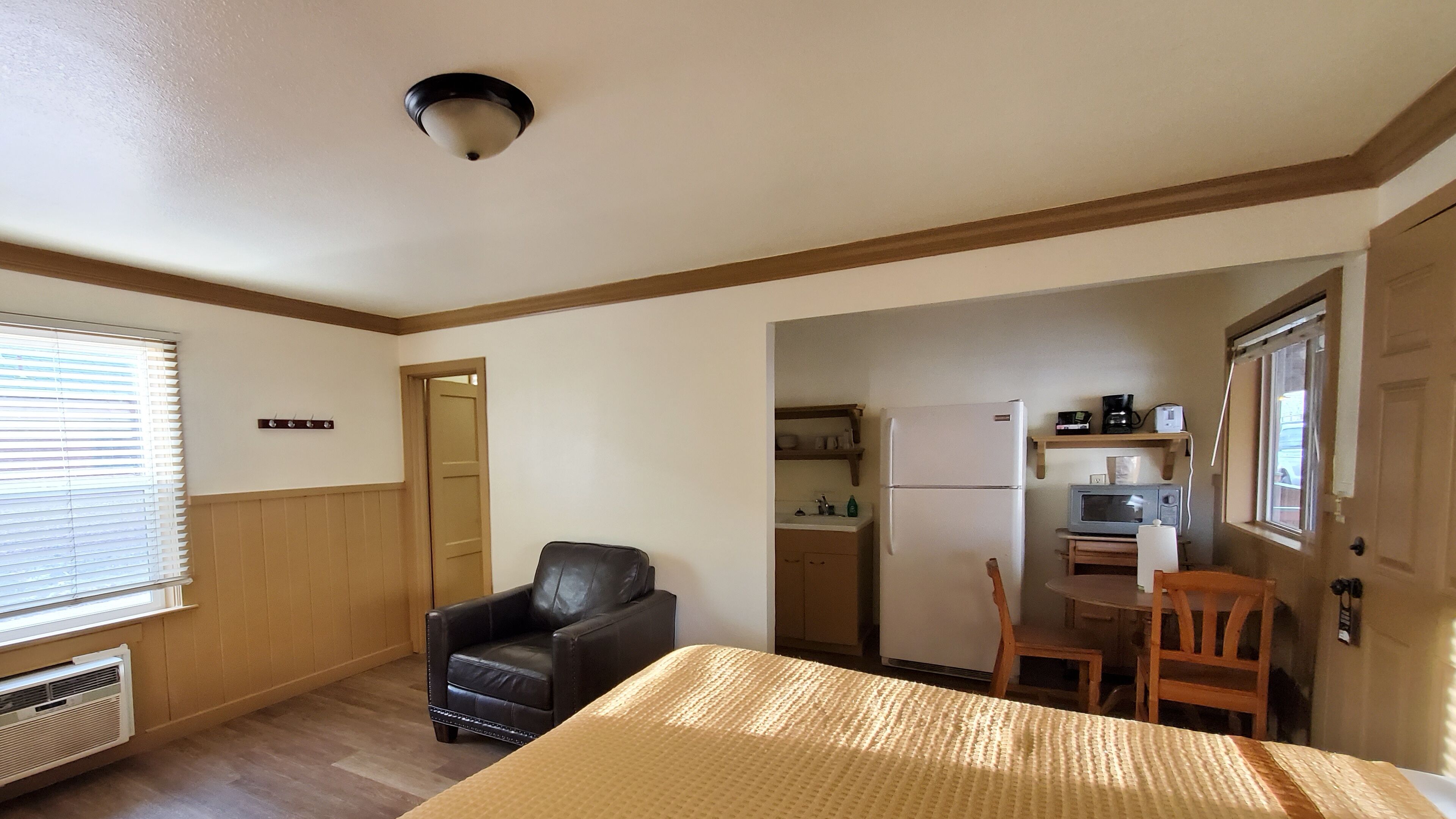 A compact room with a bed, a chair, a desk, a kitchenette, and a door leading to another room.