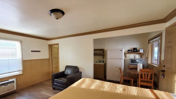 Quarto pequeno com cama, cadeira, escrivaninha, cozinha compacta e porta que dá para outro espaço.