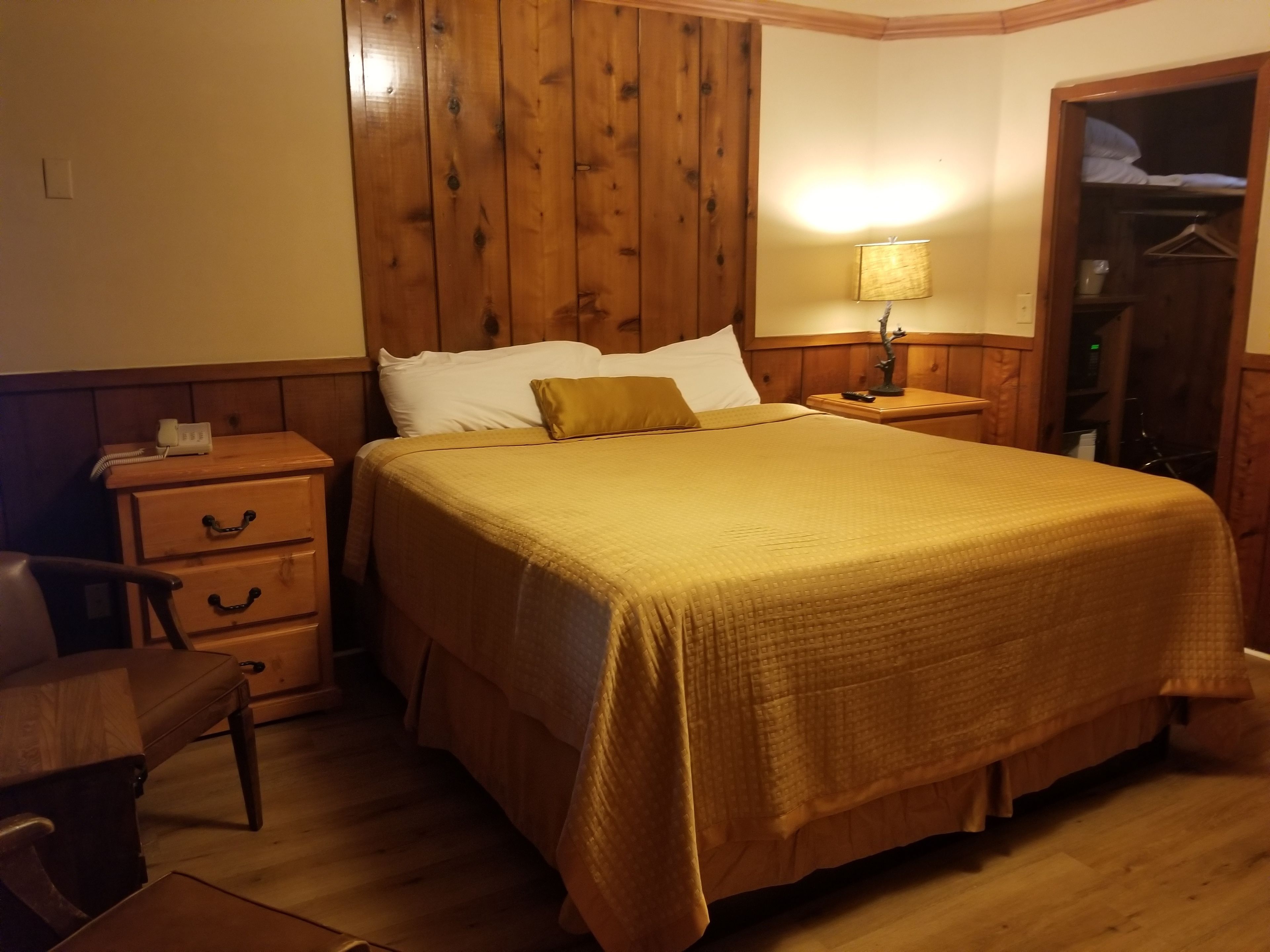 A neatly arranged bedroom with a wooden headboard, a bed with a yellow bedspread, a bedside table with a lamp, a wooden nightstand, and a chair.