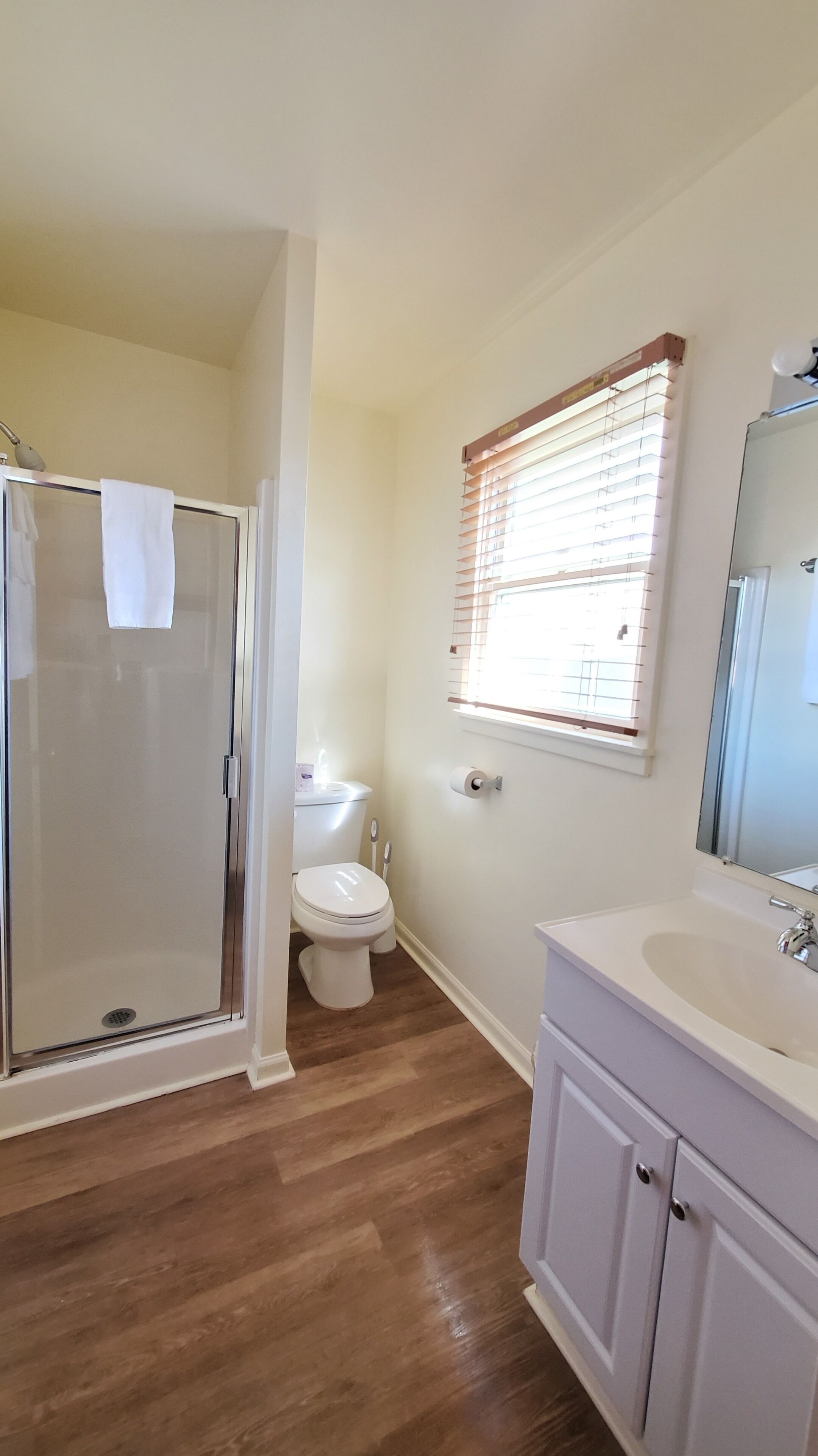 A bathroom with a glass shower enclosure, a toilet, a sink with a mirror, and a window with blinds.