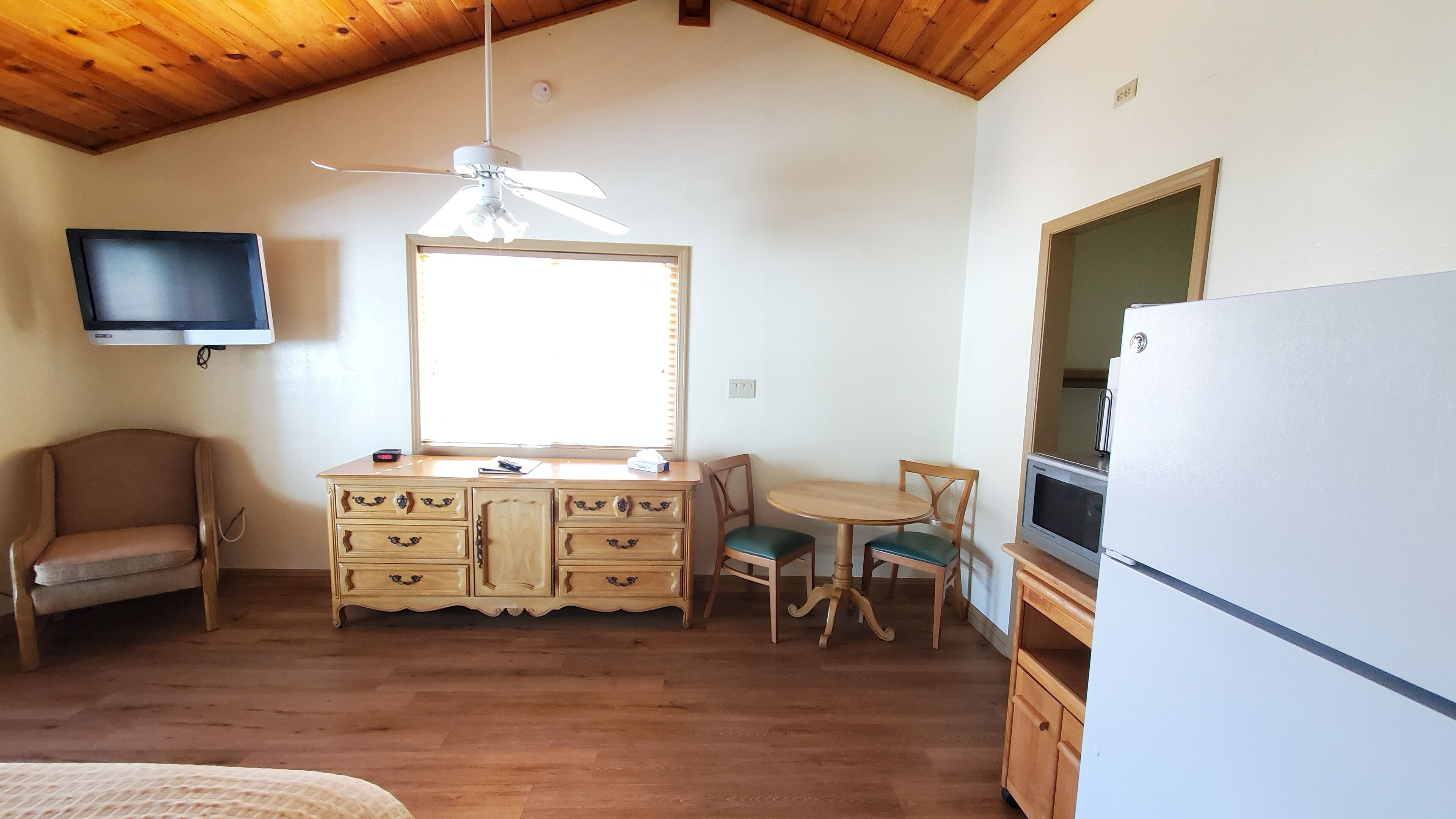 A compact room with a wooden ceiling, a ceiling fan, a television mounted on the wall, a wooden cabinet with drawers, a small dining table with two chairs, and a refrigerator.