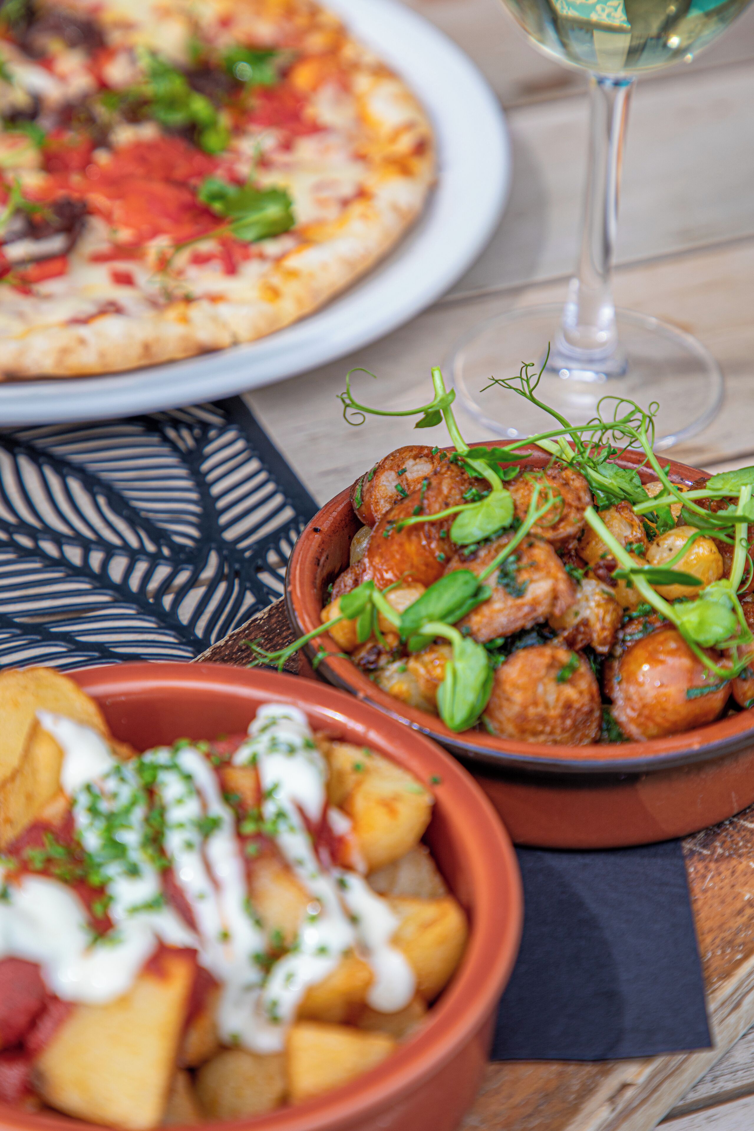 A dining table with a plate of pizza, a bowl of meatballs with greens, and a bowl of potatoes with sauce.