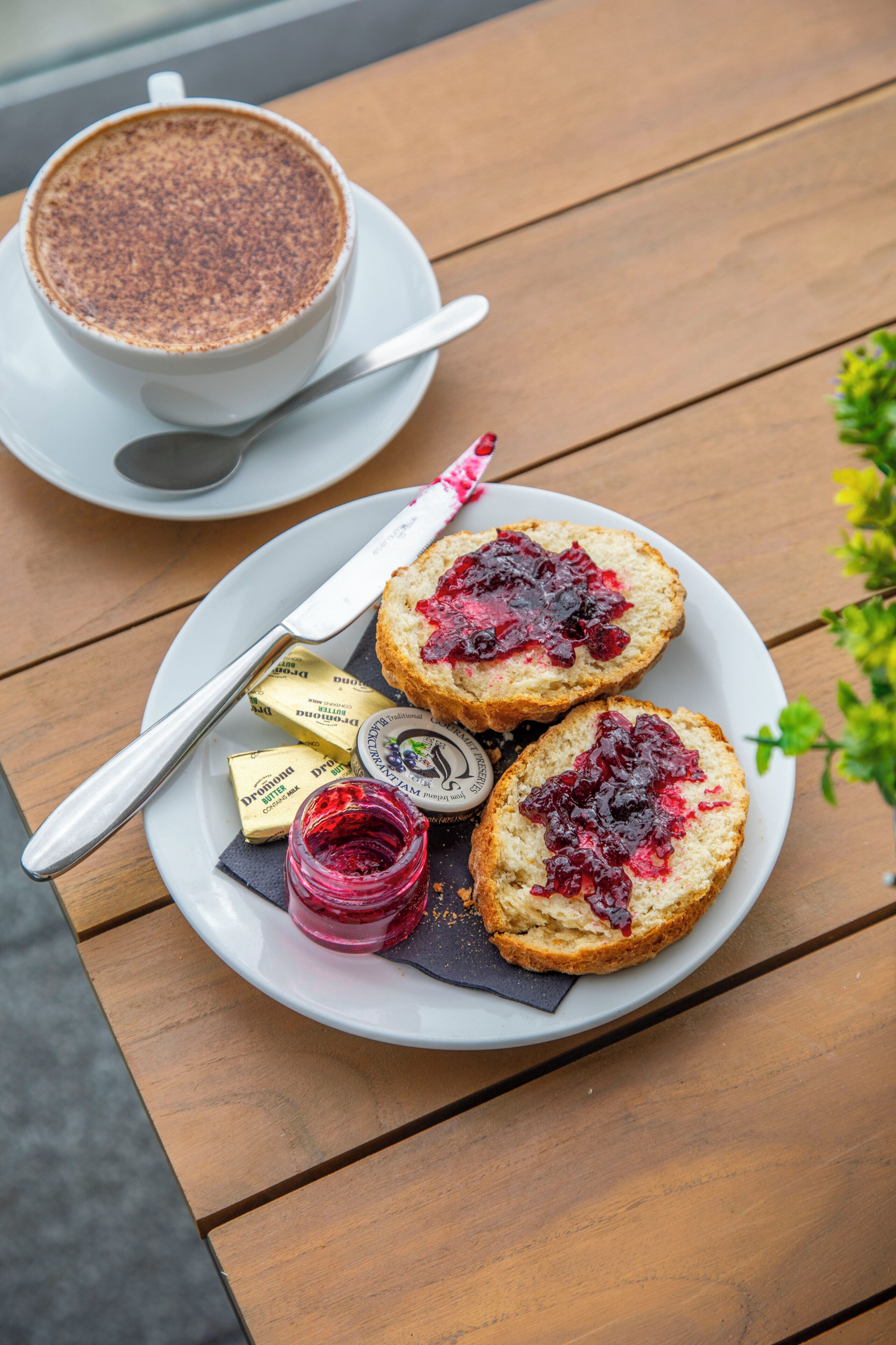 A plate with two scones topped with jam, a jar of jam, a knife, and a cup of coffee on a wooden table.