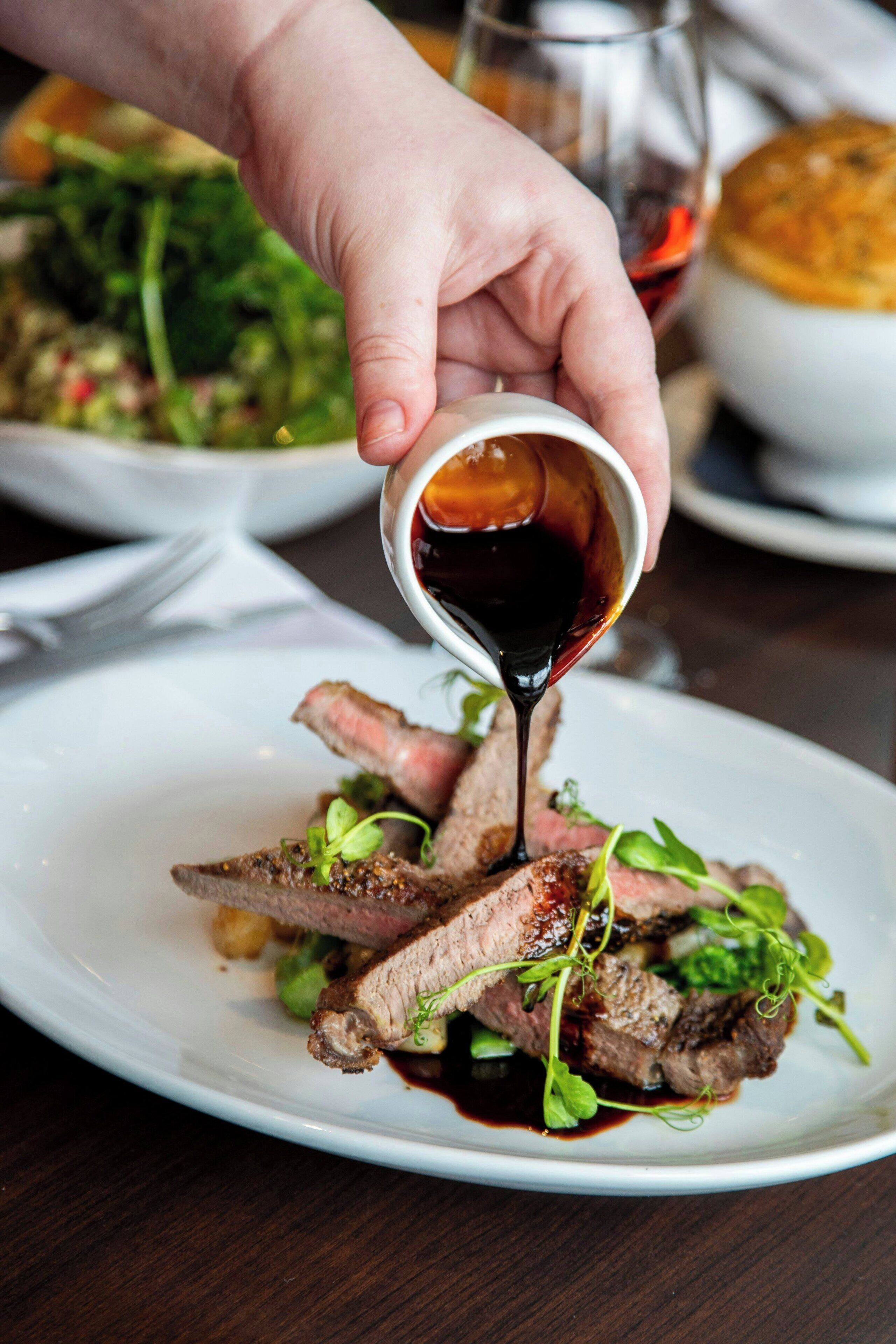A hand pouring sauce over a plate of steak with greens.