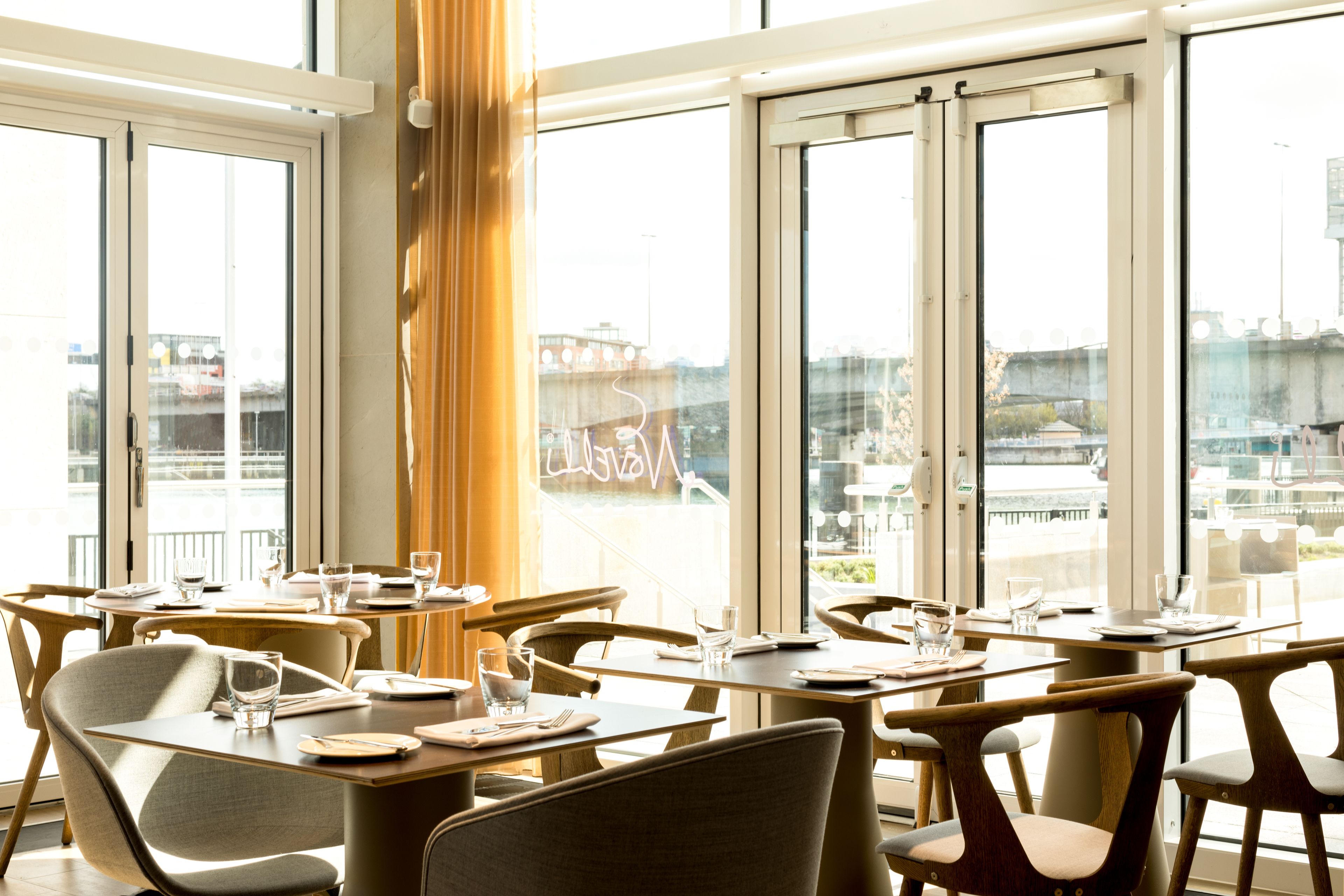 A modern dining area with multiple tables and chairs, large windows offering a view of the cityscape.