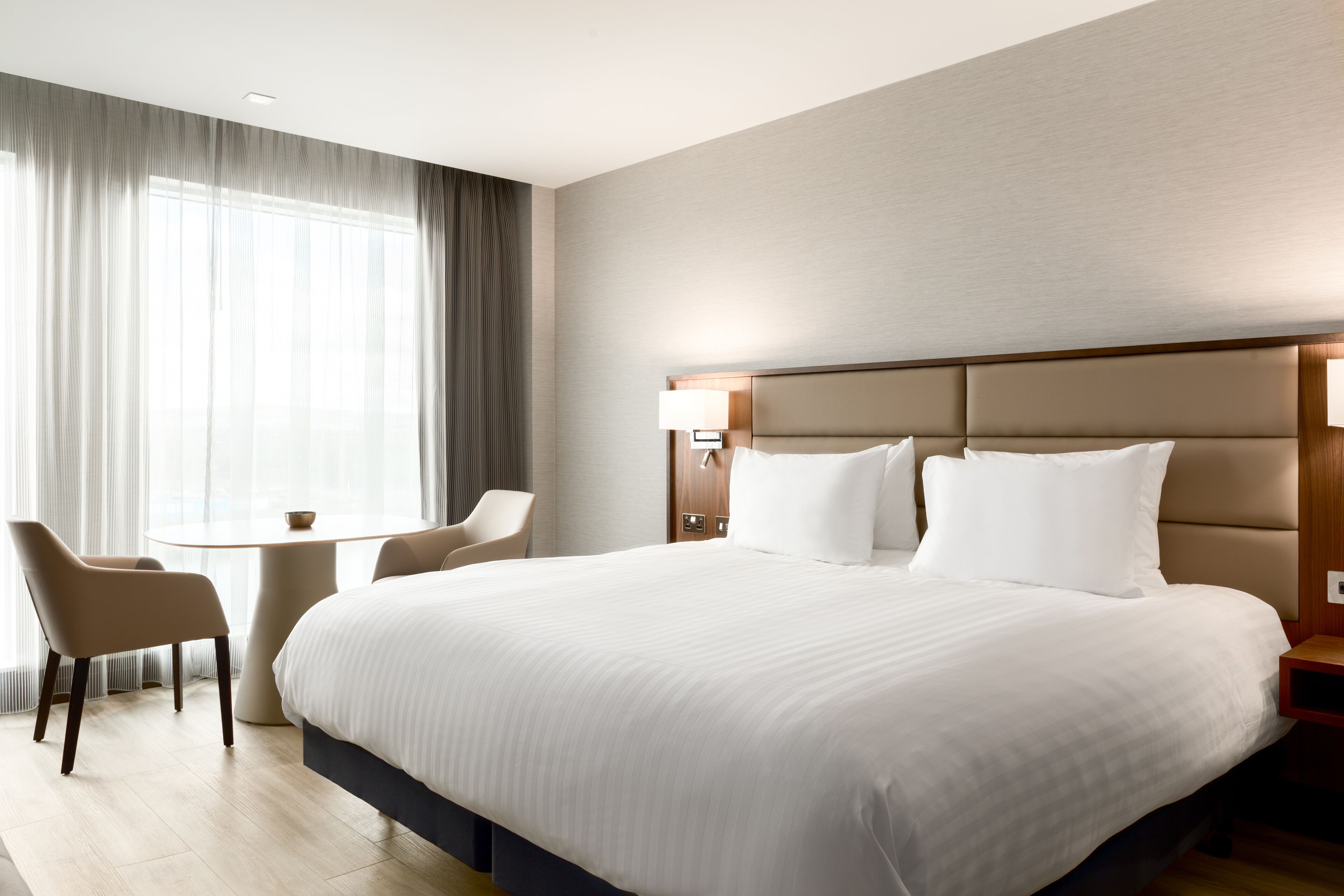 A hotel room with a large bed, a bedside table, a chair, and a window with curtains.